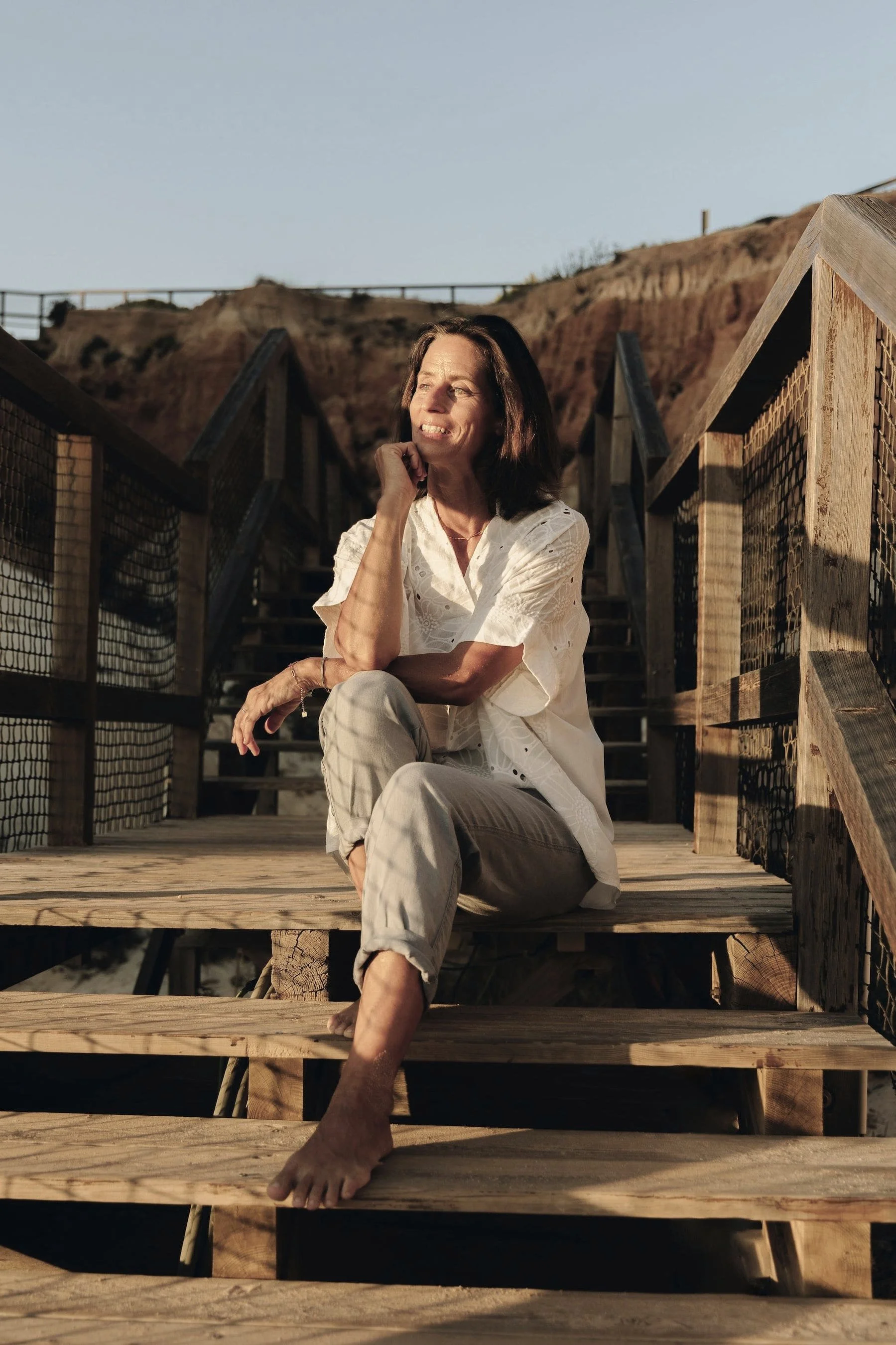 Eine Frau sitzt entspannt auf einer hölzernen Treppe im Freien bei Sonnenuntergang, mit Blick auf eine felsige Landschaft im Hintergrund.
