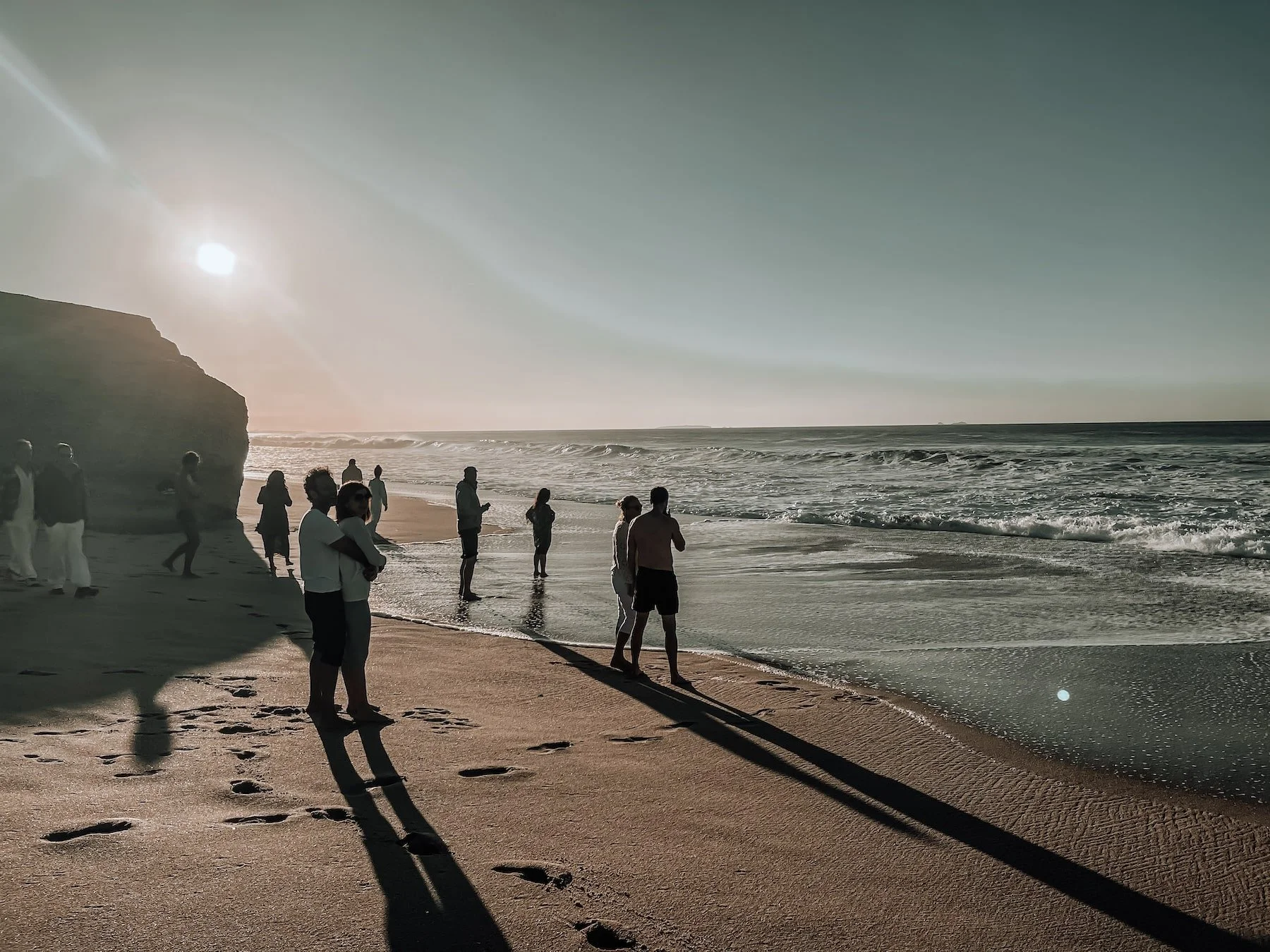 Menschen stehen bei tief stehender Sonne an einem Sandstrand mit Felsen und welligem Meer.