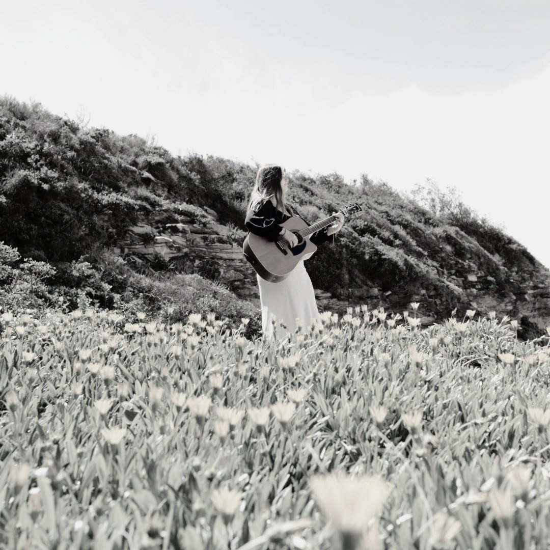 A woman playing guitar in a field of flowers, with hills in the background, in black and white.
