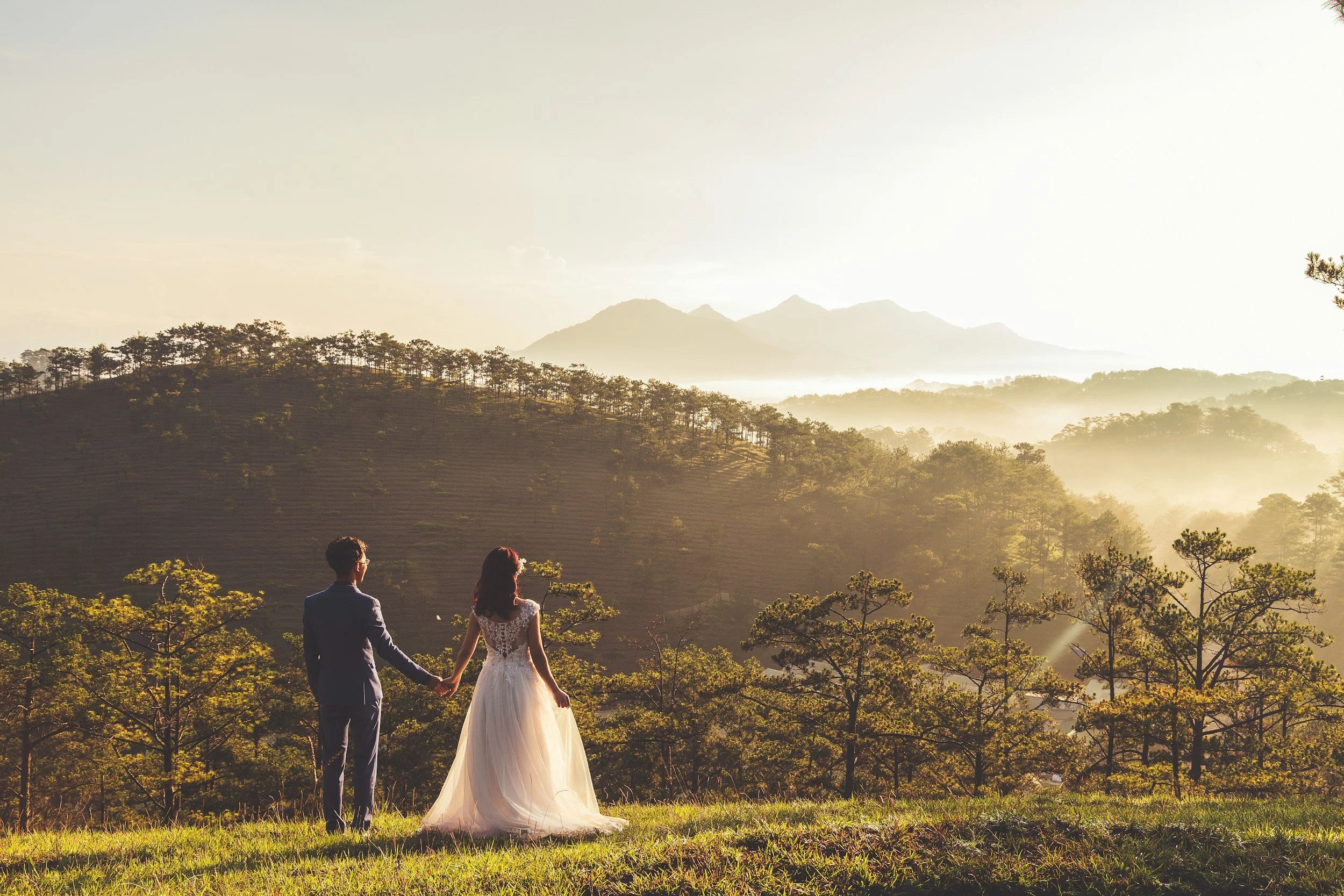 A bride and groom holding hands, standing on a grassy hilltop with a mountain landscape in the background during sunrise or sunset.