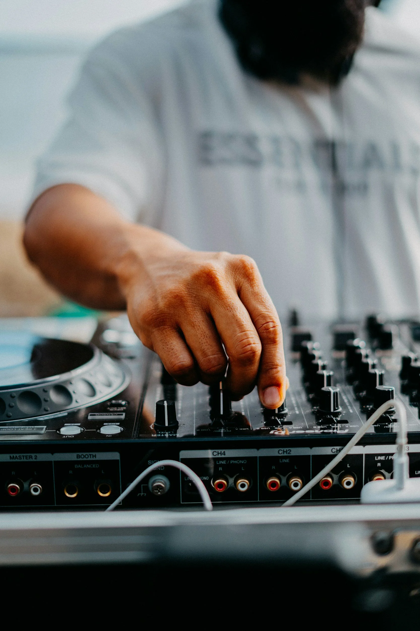 DJs performing at an outdoor event, with one wearing sunglasses and headphones, operating a DJ mixer, and the other wearing a cap and headphones, raising his hand in the background.