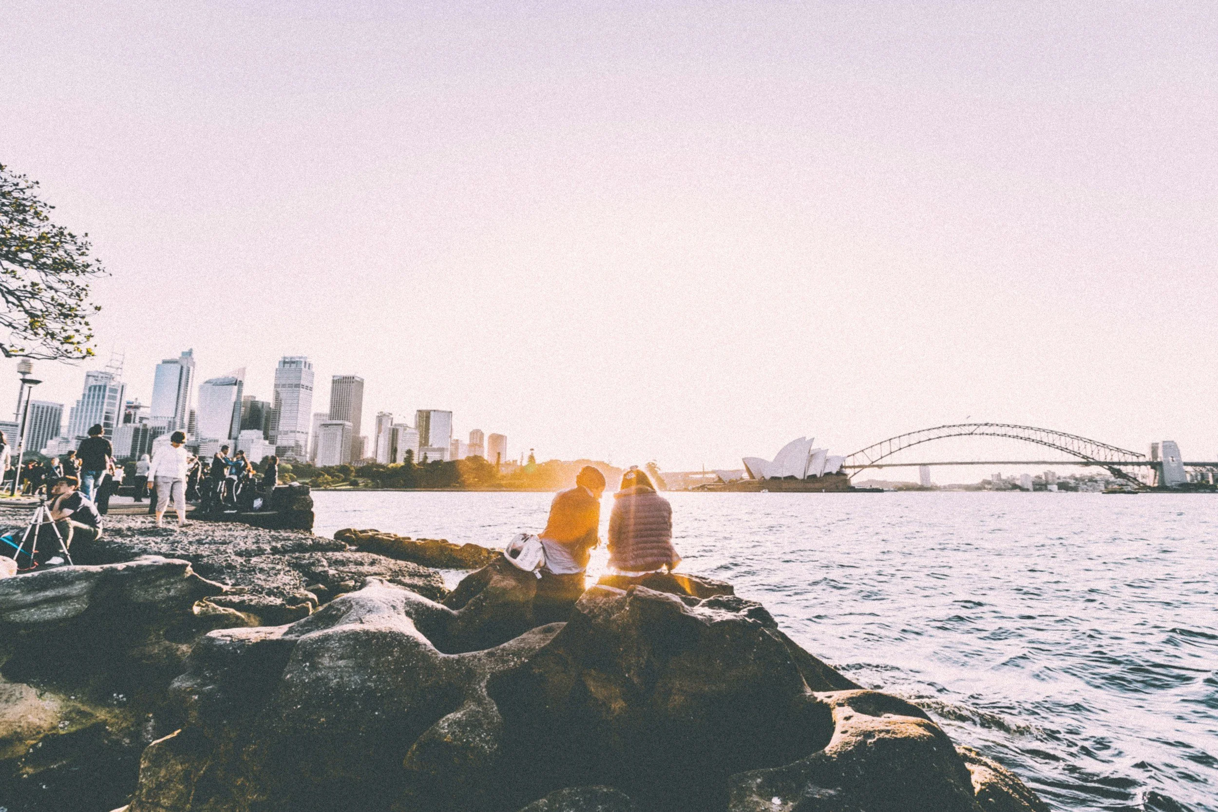 Two people sitting on rocks beside Sydney Harbour, with the city skyline and Sydney Opera House in the background, during sunset.