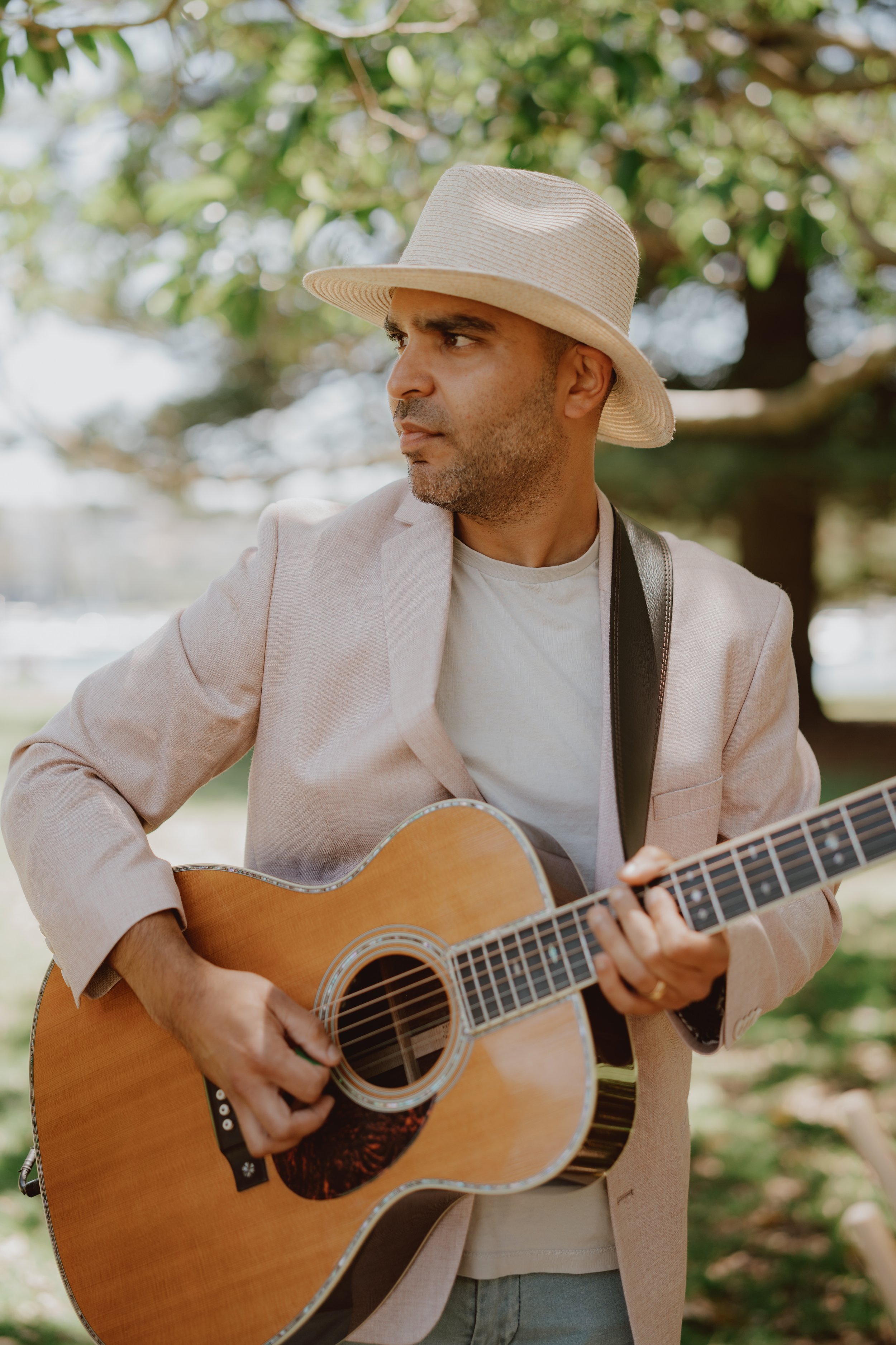 Man playing guitar outdoors wearing a hat and light-colored blazer.