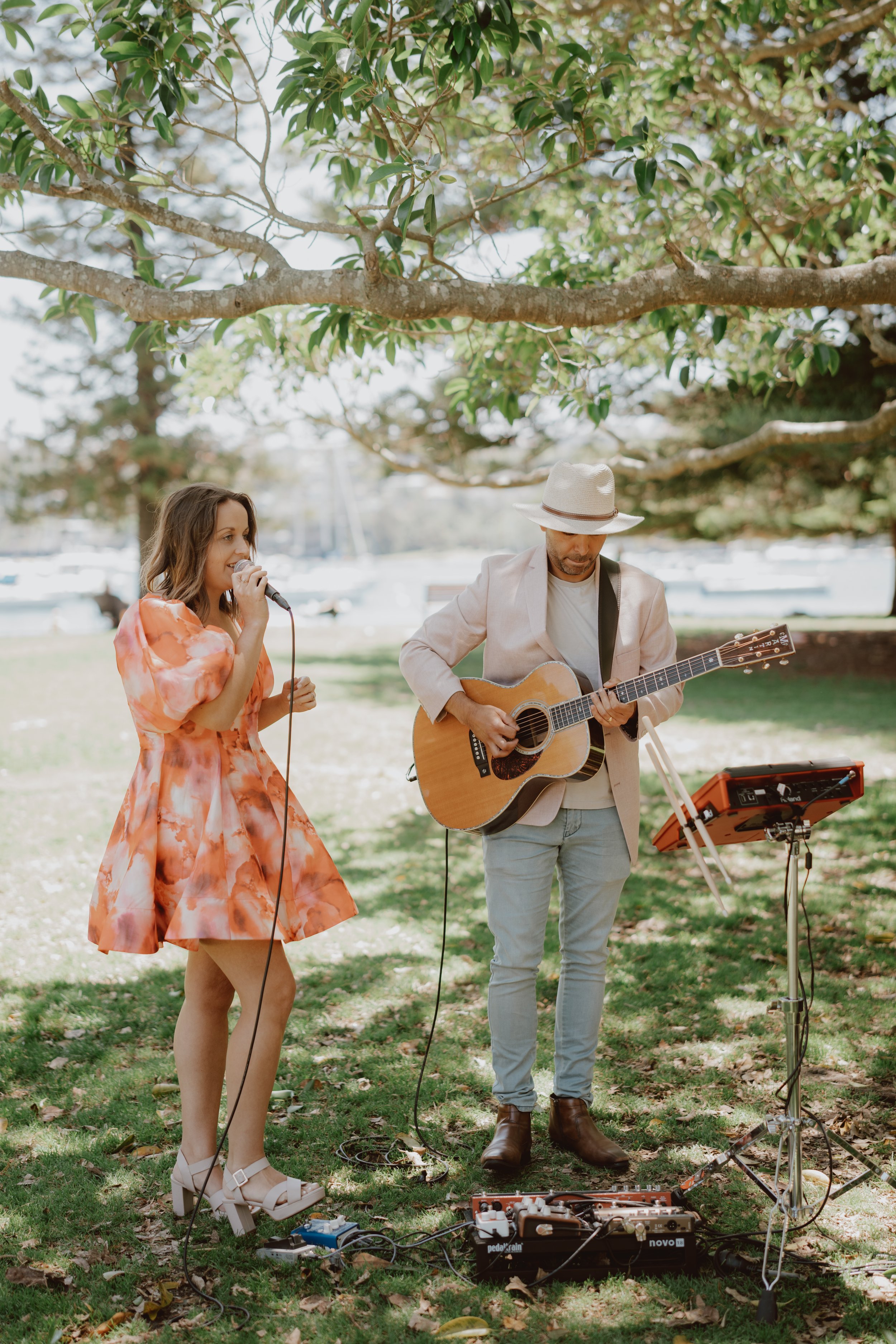 Two musicians performing outdoors; a woman in an orange dress sings into a microphone, and a man wearing a hat plays an acoustic guitar. They are under a tree on a grassy area with equipment around their feet.