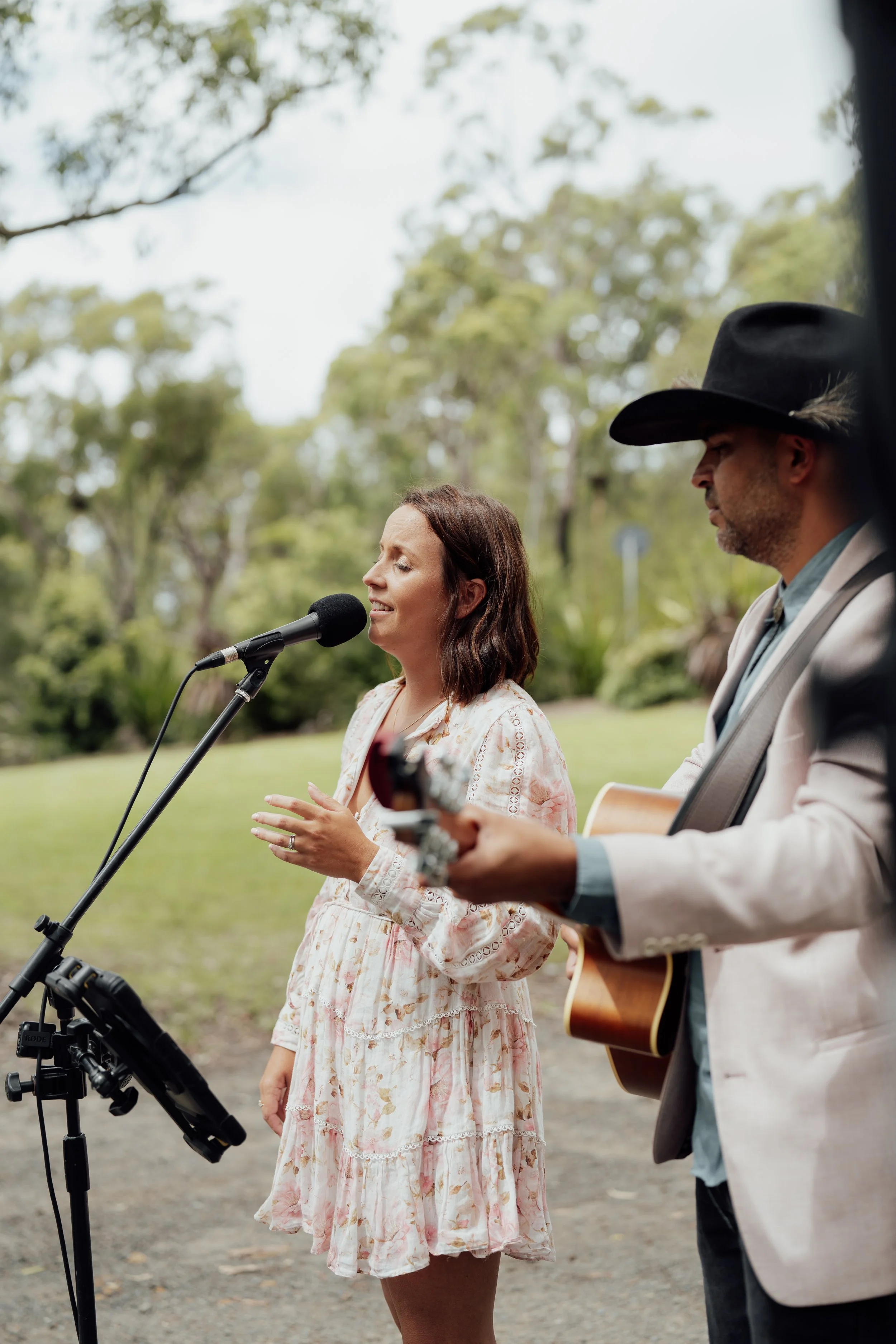 A woman singing into a microphone and a man playing guitar outdoors in a park with green trees.