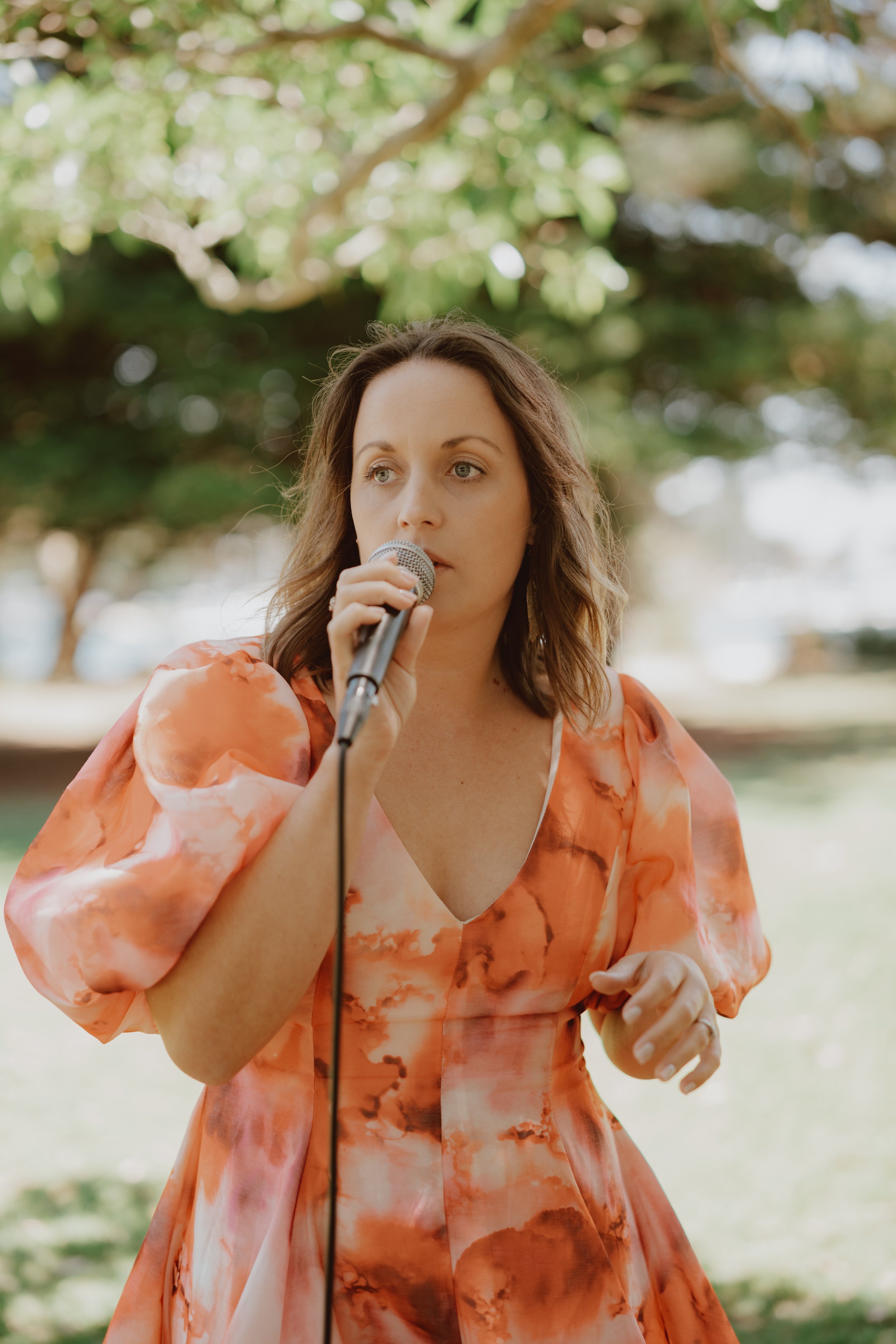 Woman singing outdoors with a microphone, wearing an orange patterned dress.