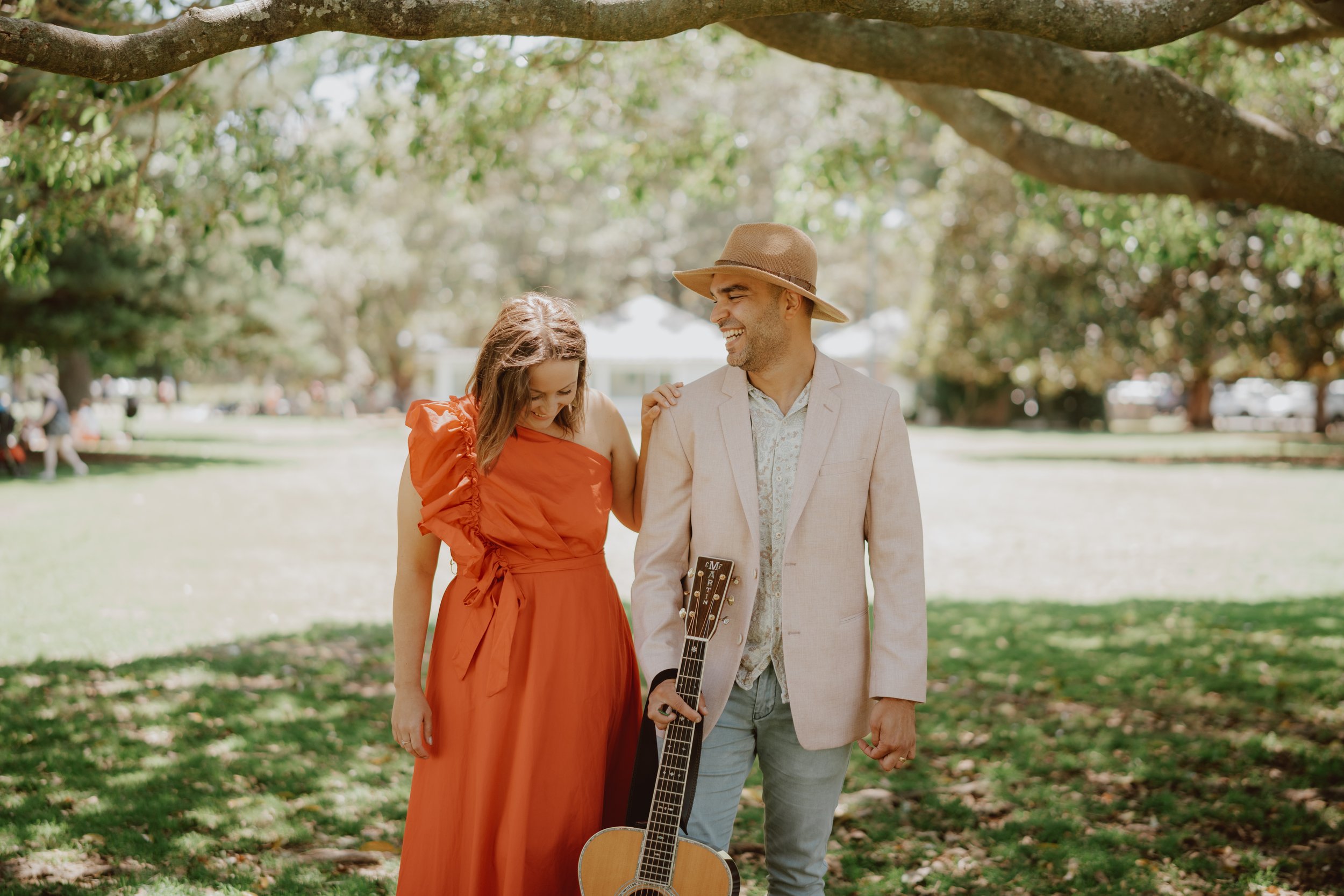 A man and a woman walking in a park, with the man holding a guitar. The woman is wearing a red dress and the man is dressed in a light blazer and hat.