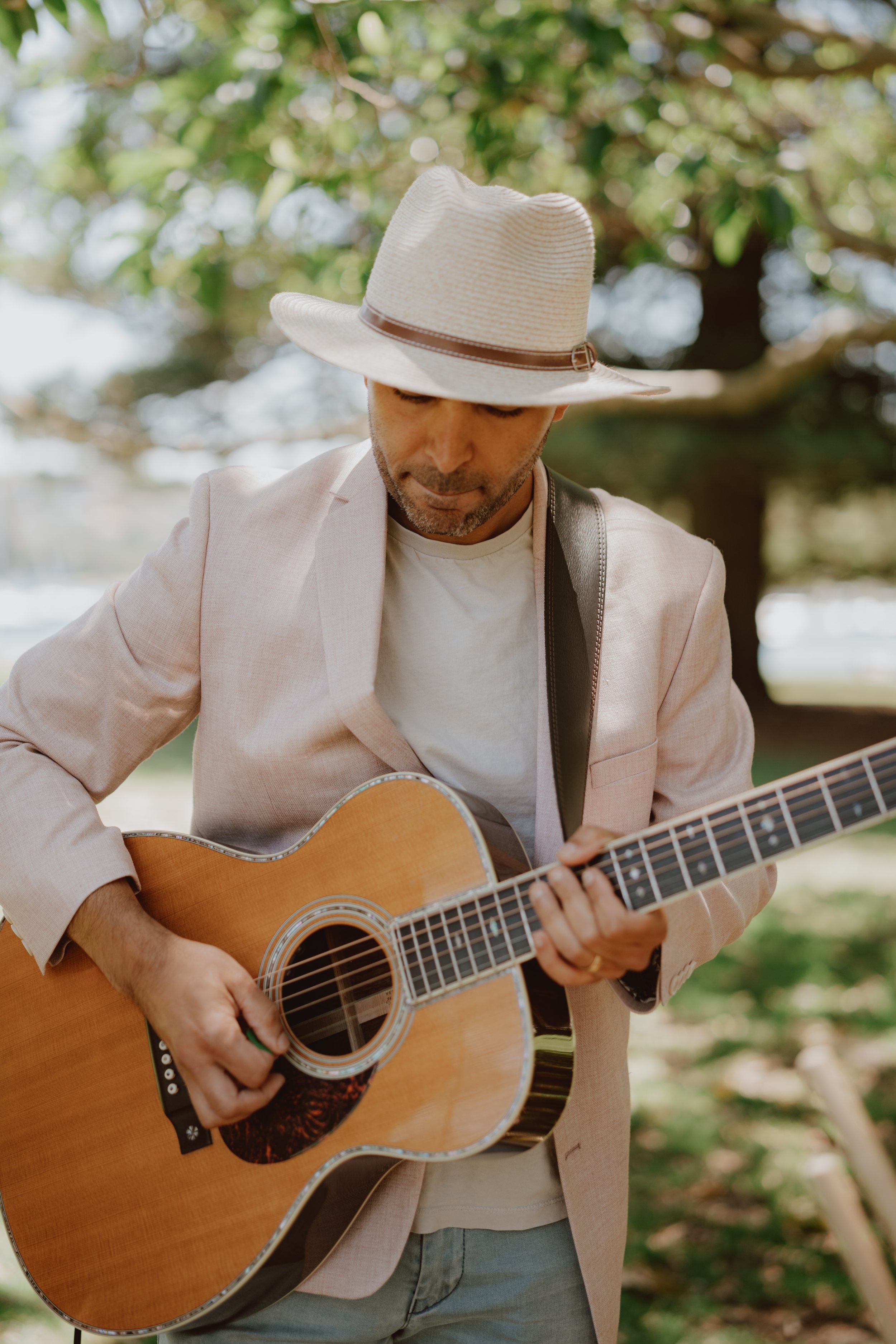 Man playing acoustic guitar outdoors wearing a beige jacket and straw hat.