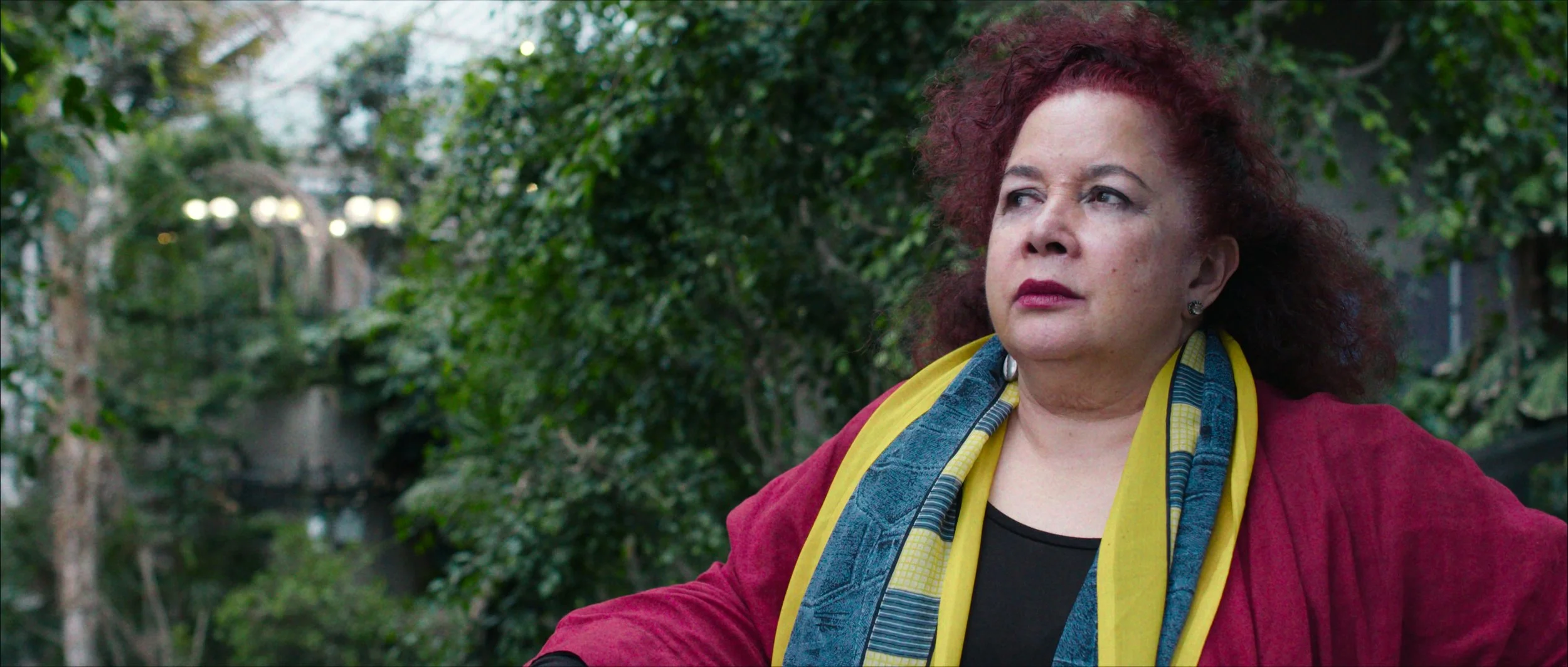 A woman with red curly hair looks off into the distance of the Barbican Conservatory, GRETA