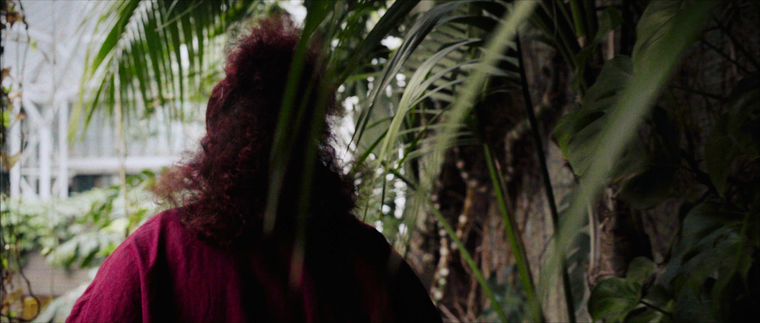 A woman with red curly hair walks across a gangway in the Barbican Conservatory, GRETA