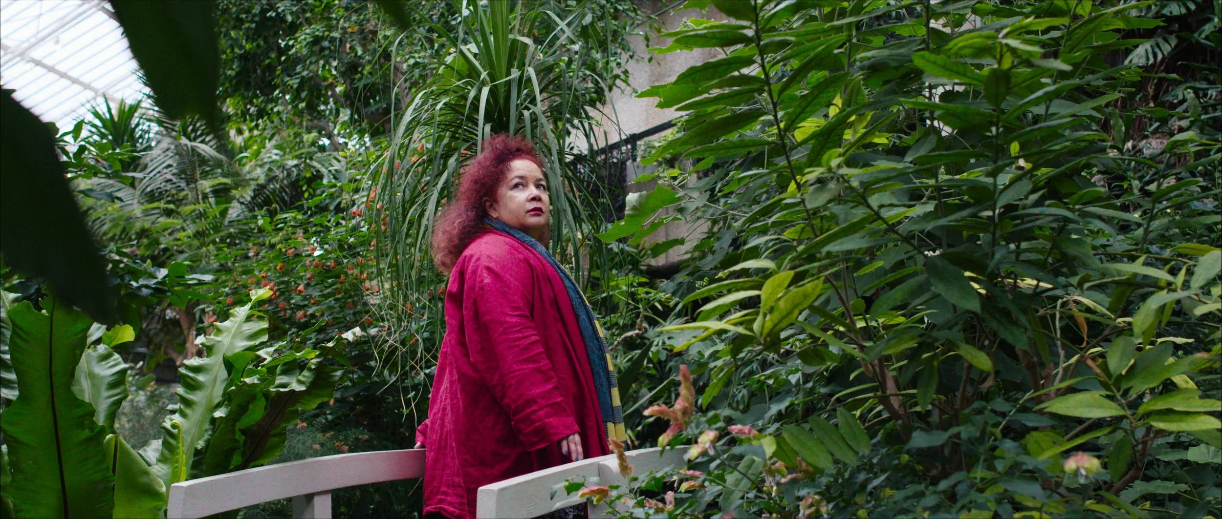 A woman with red curly hair walks across a bridge in the Barbican Conservatory, GRETA