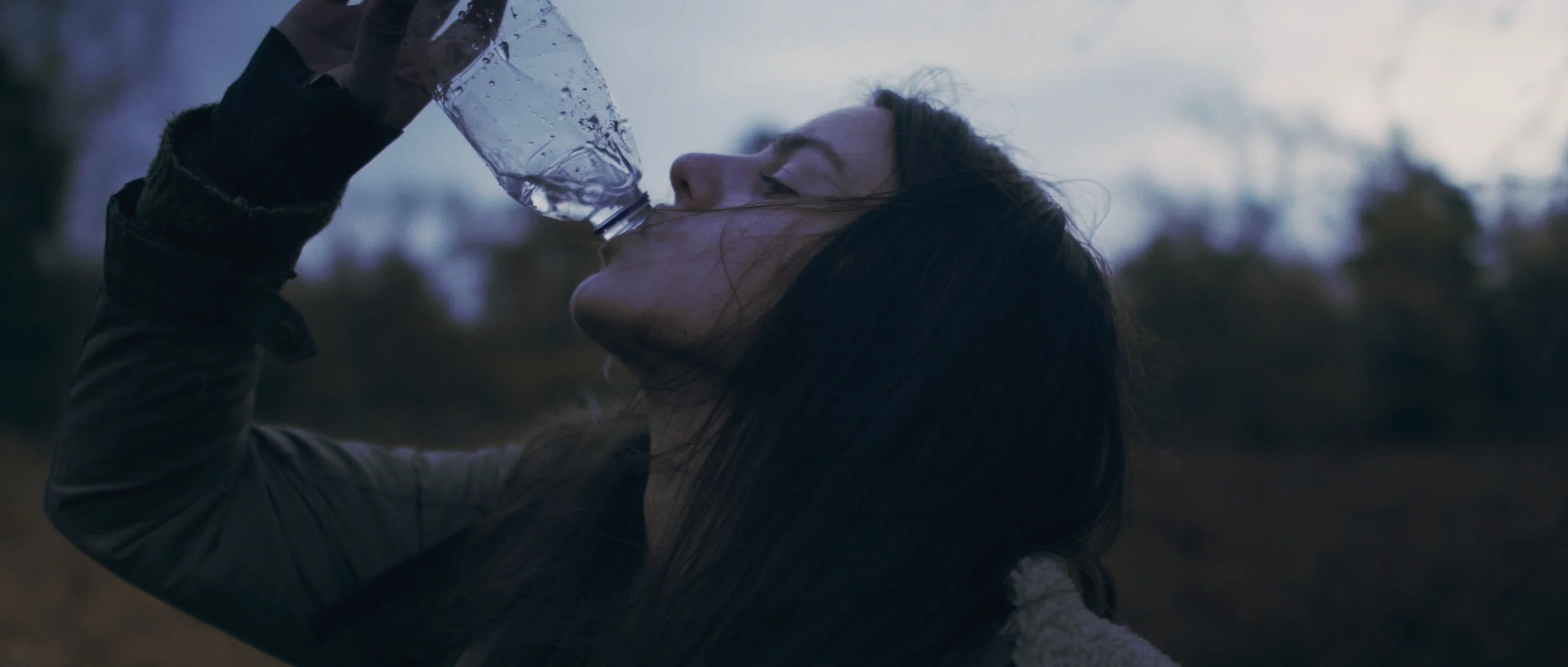 A woman drinks from a near empty water bottle at dawn in the forest, Flying Blind