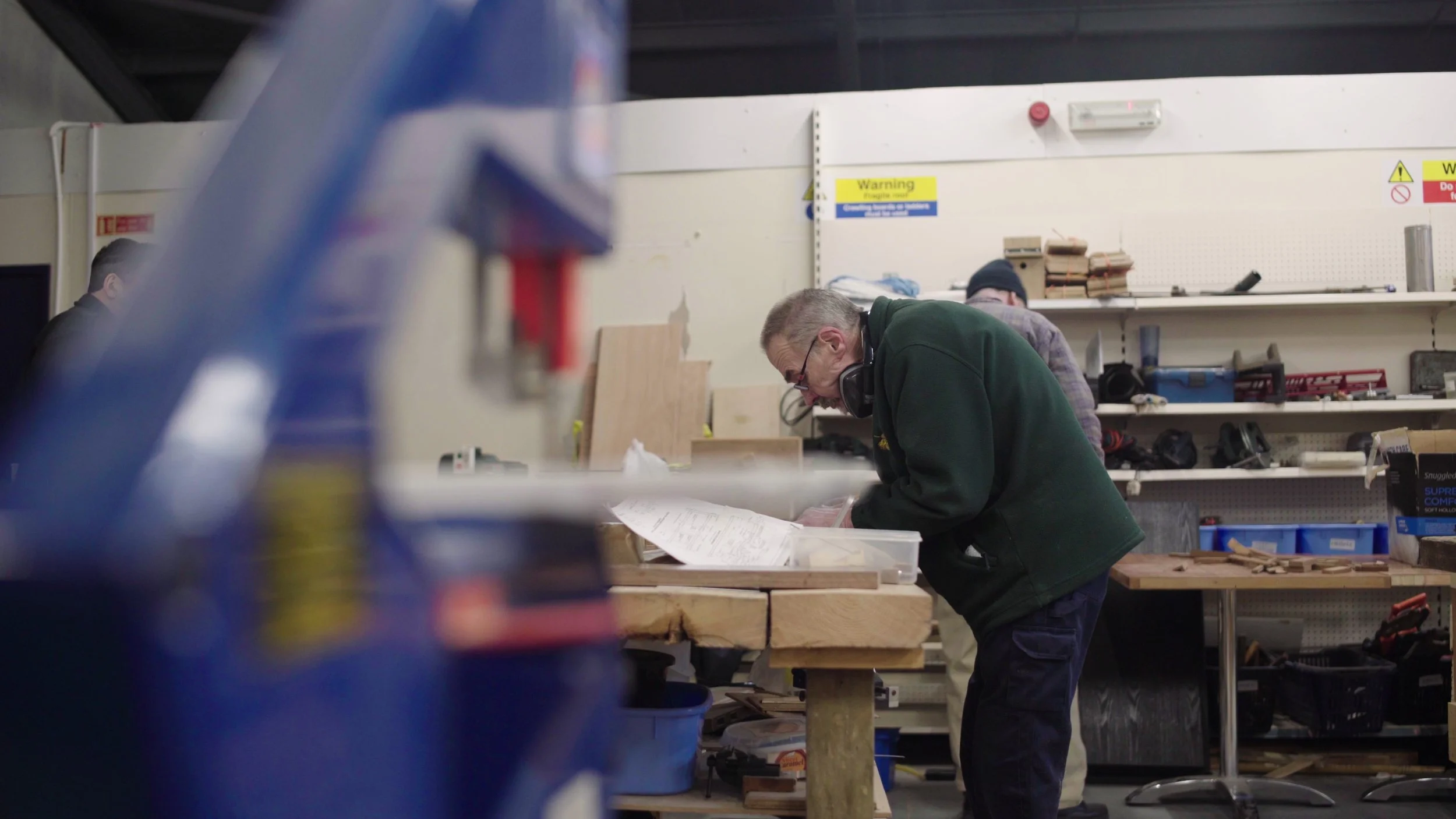 A man in a green jacket works on a wood project as a blue machine is in the foreground, Men's Shed