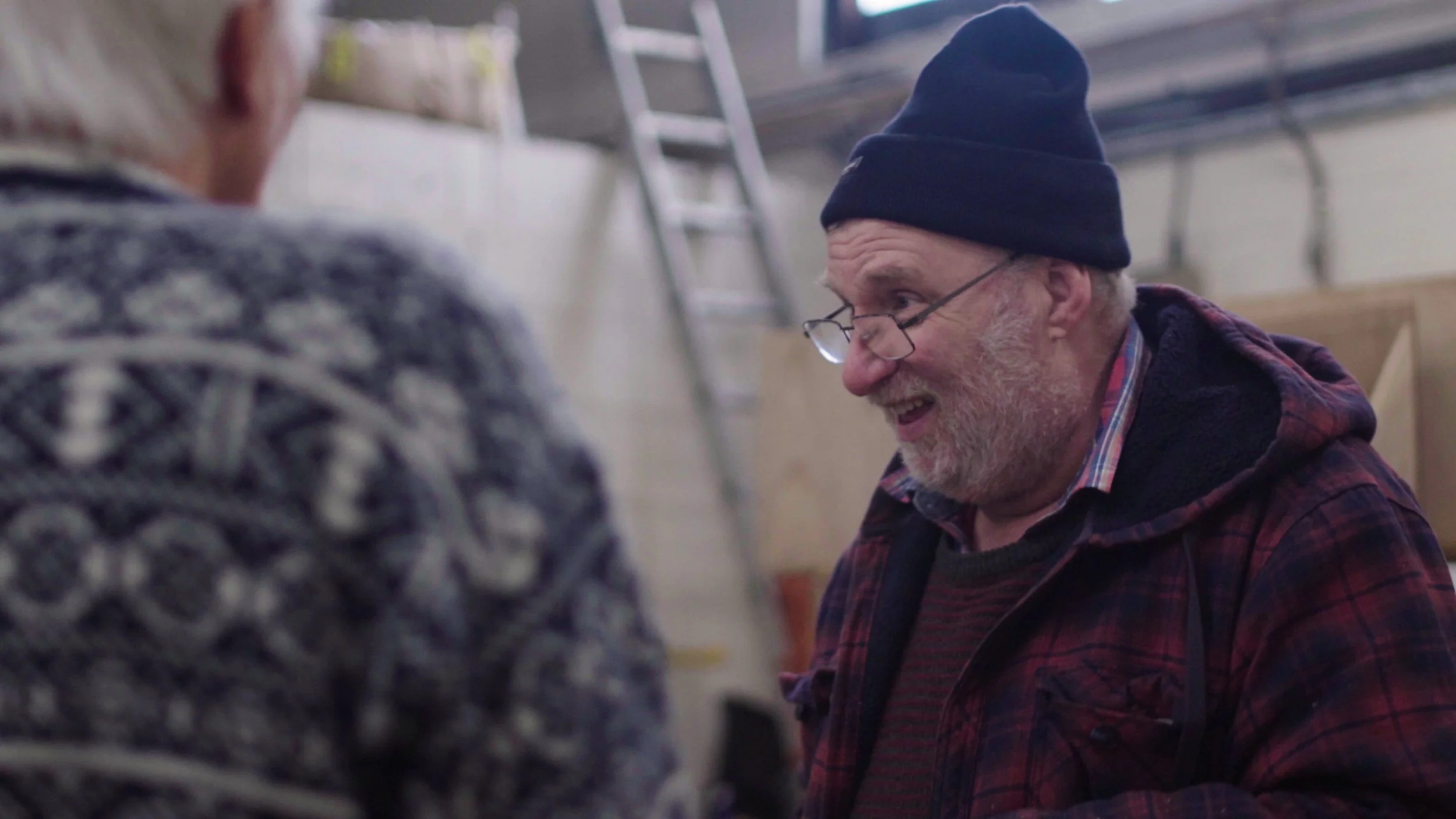 A man in a blue beanie hat smiles in the workshop, Men's Shed