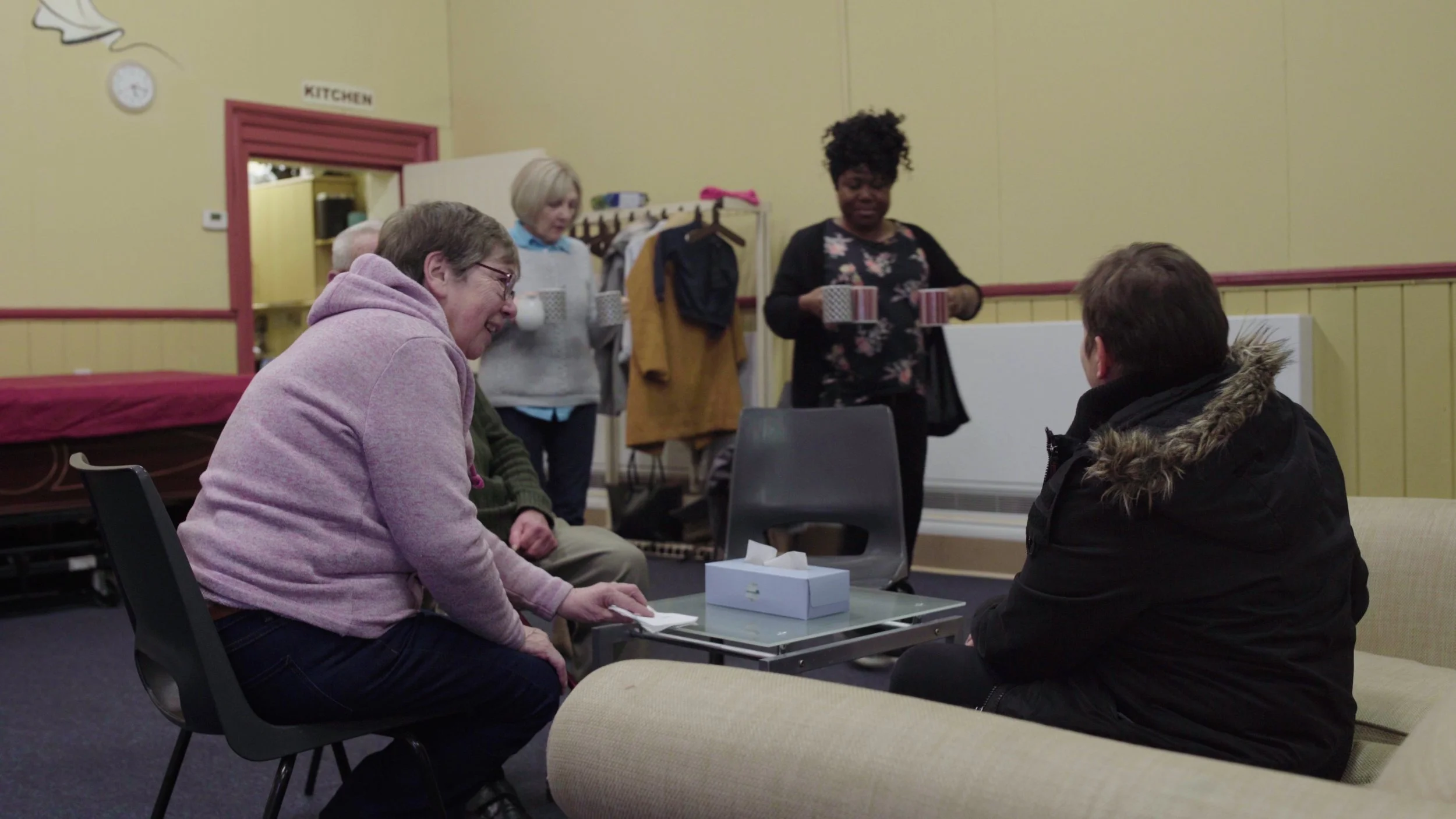Two women bring hot drinks to the group while a few people sit around a table, Warwick Bereavement Group