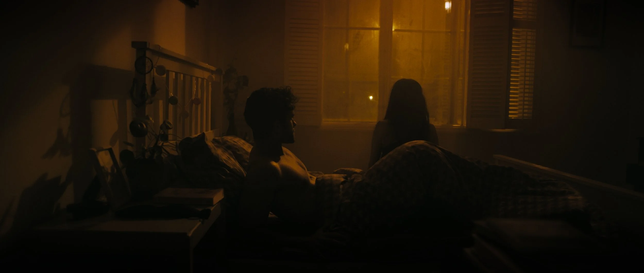 A man lays in bed as a woman sits on the edge of the bed. Both are silhouetted against orange coloured streetlights, Night Whispers