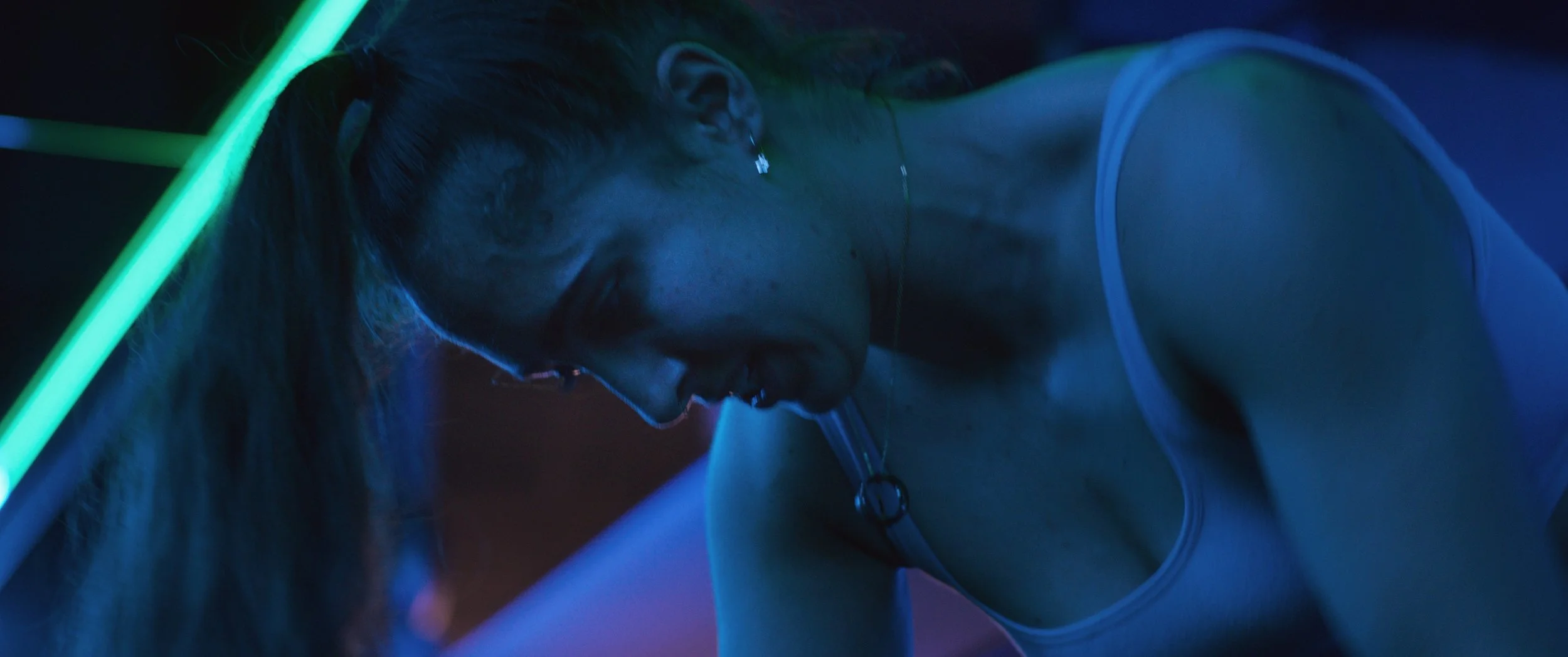 Hajnalka Balazs (Hajni) hunches over in a dimly lit gym with green and blue lighting while doing one-armed rows, Easy