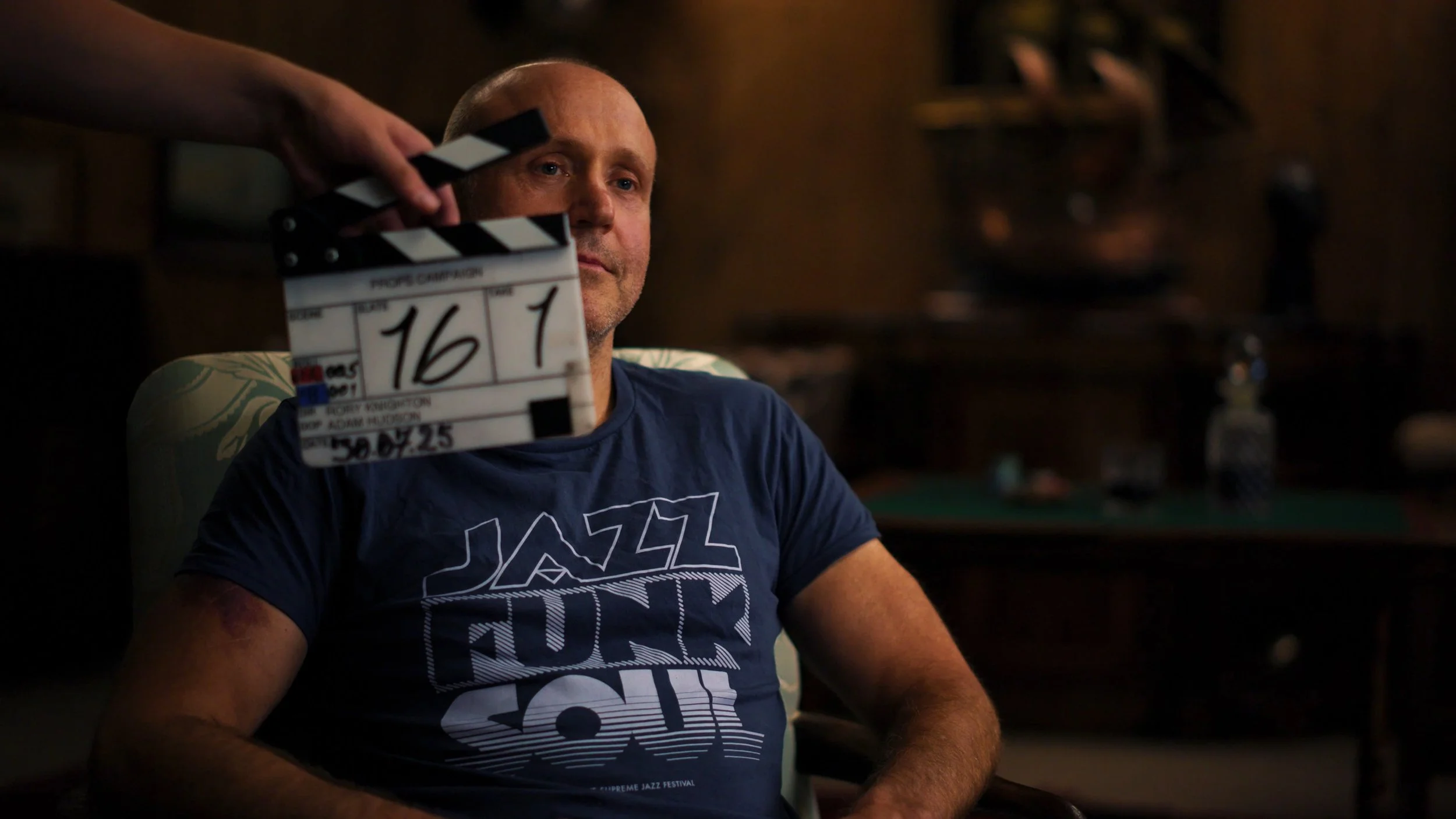 Spencer Jordan sits in a chair in a prop warehouse with a film slate in front of his face, The Secret Life of Props