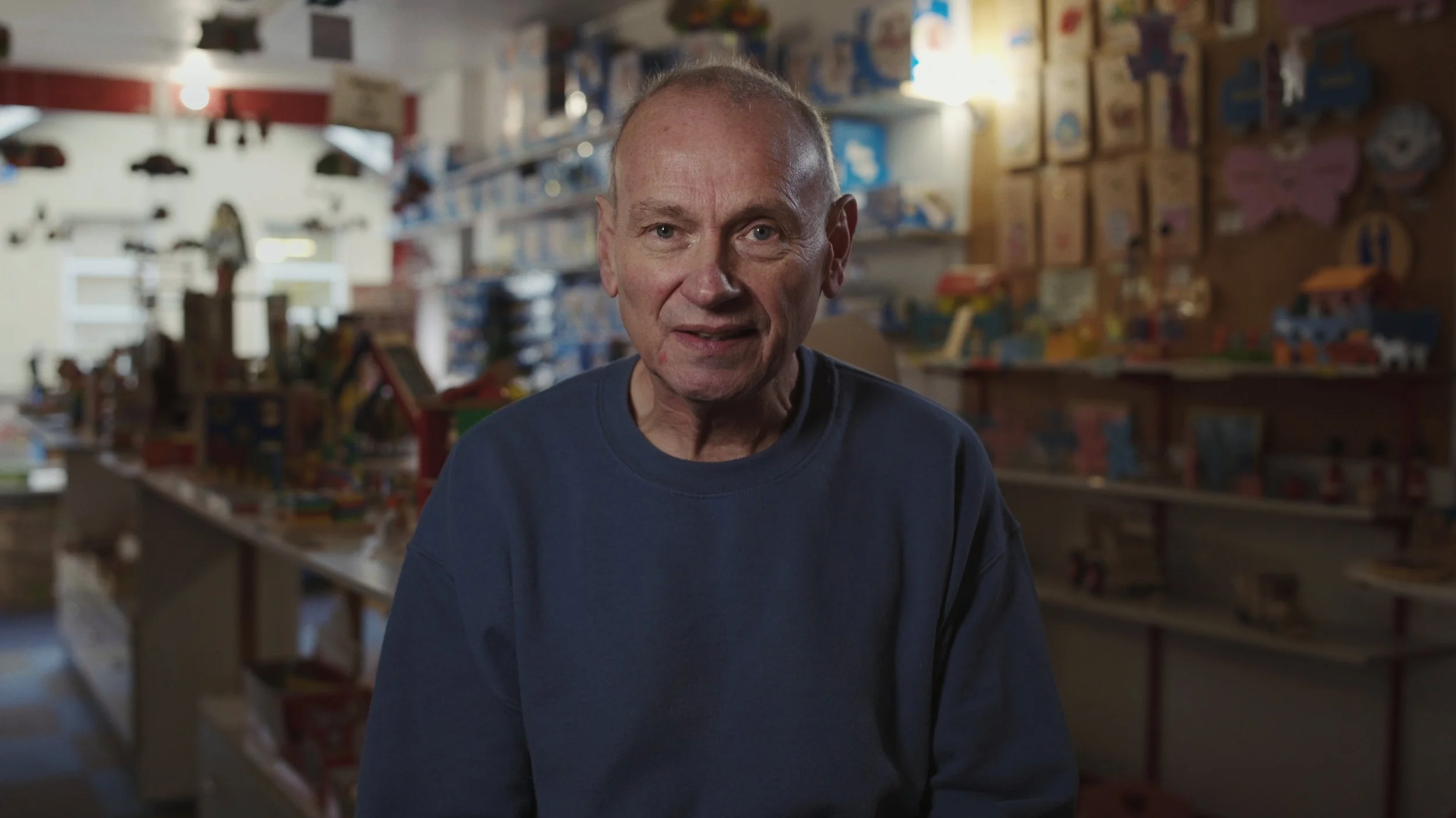 An elderly man sits in a wooden toyshop while looking into camera, The Last Toymaker