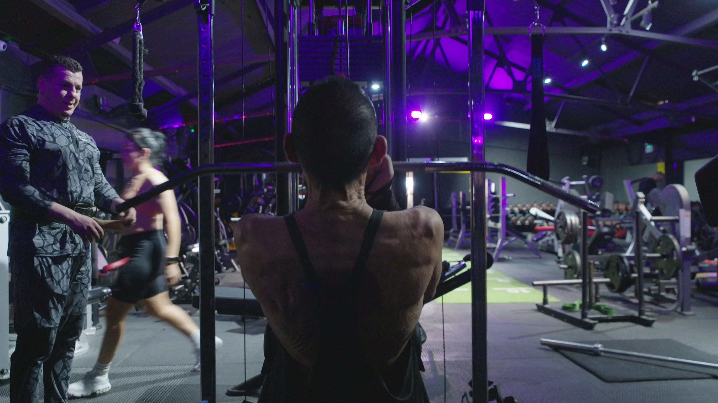 An elderly man in a dimly lit gym with purple lighting works out at the lat pull-down machine, King Dick