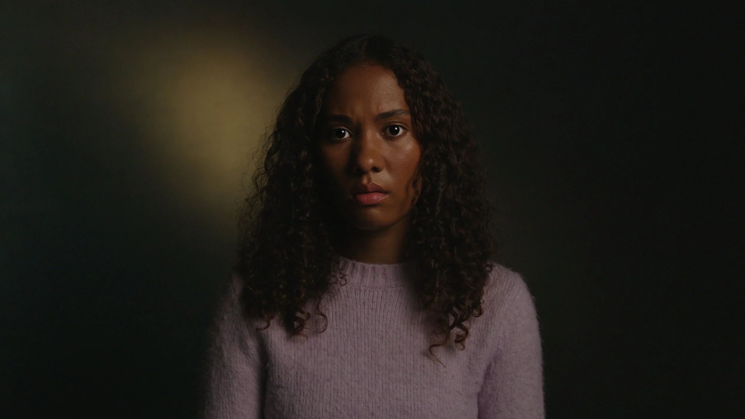 A woman with curly hair in a dark room looks into camera, It's Not Just Banter