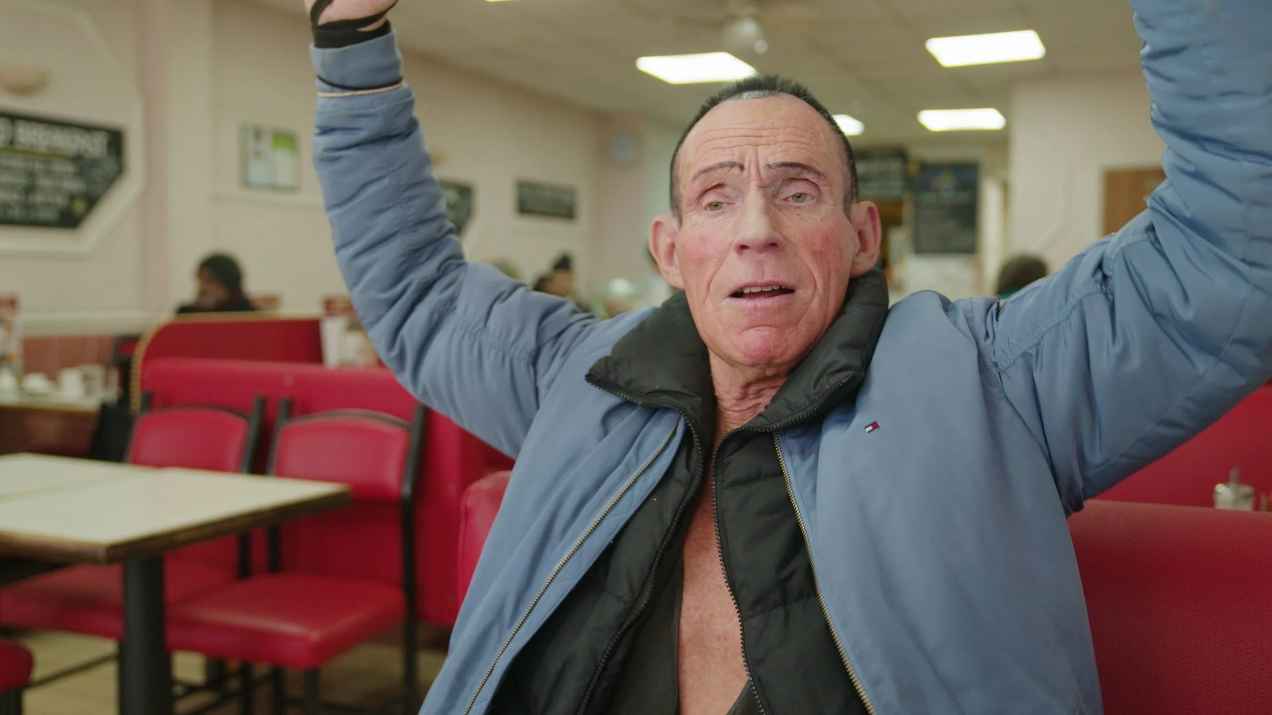 An elderly man raises his arms above his head while sitting in an old-school London café, King Dick