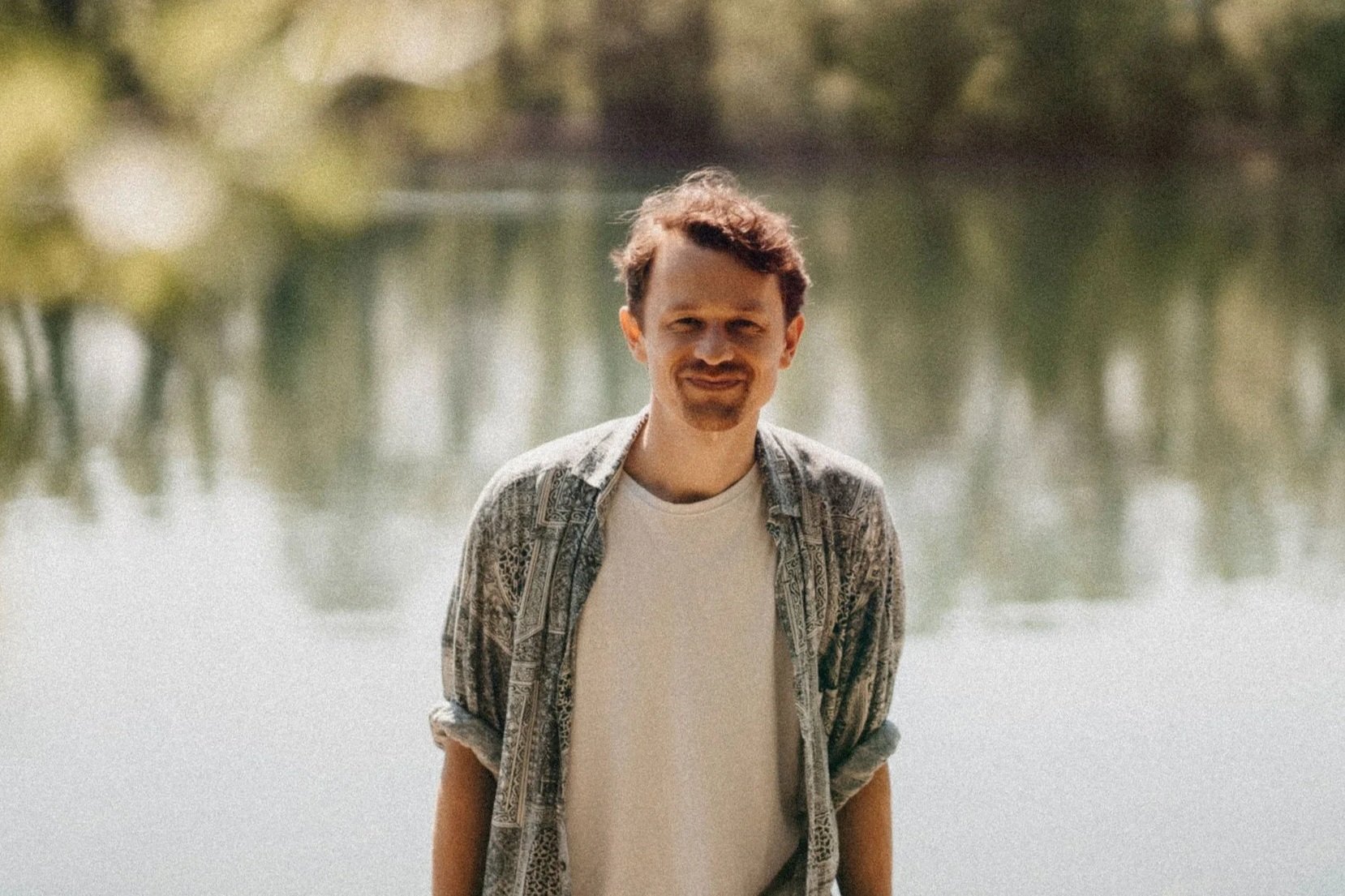 Lukas Leitinger steht am Ufer eines Sees, trägt ein T-Shirt und ein offenes Hemd, im Hintergrund sind Bäume und Wasser zu sehen.