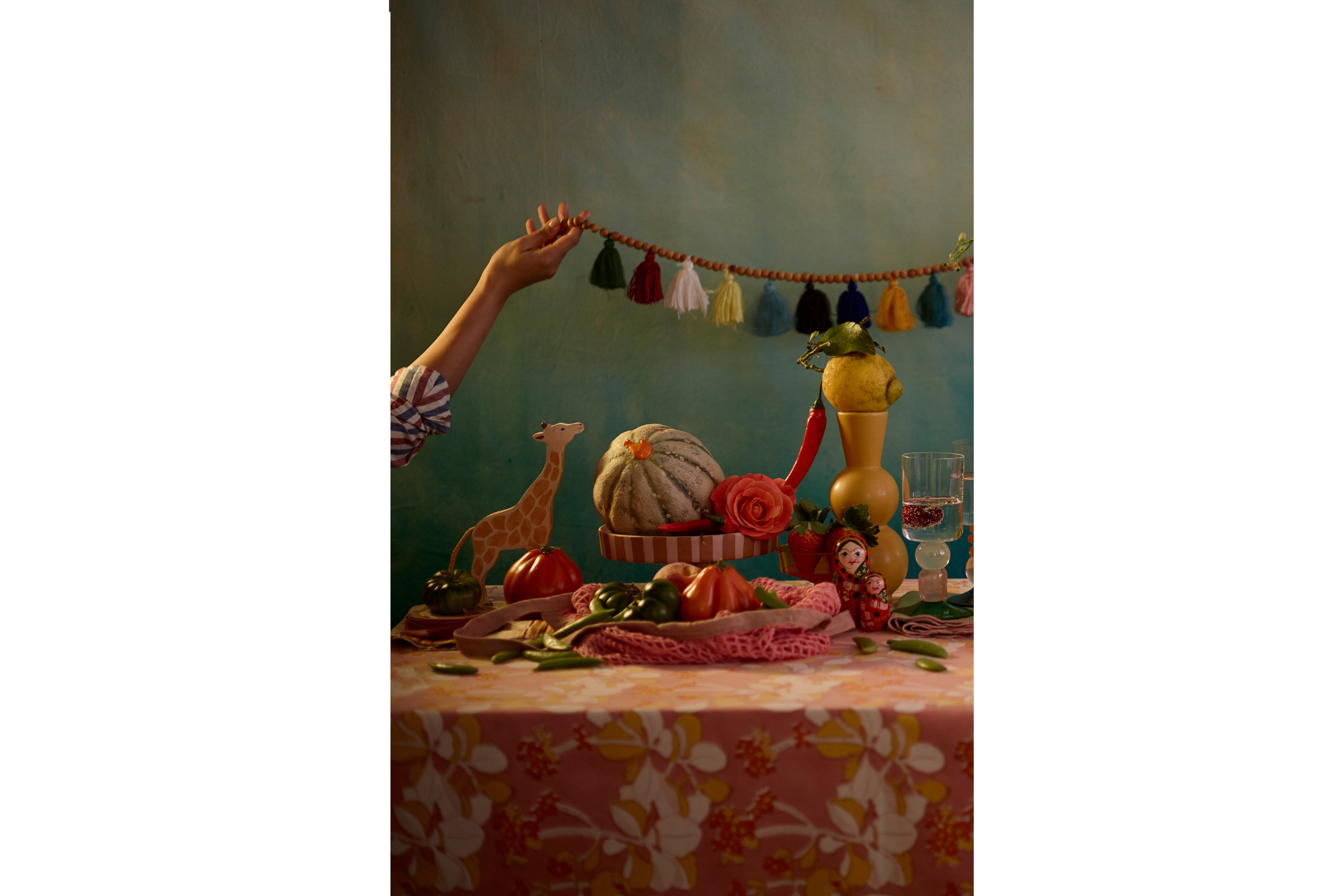 A colorful fiesta table setup with fruits, vegetables, a gourd, a plastic giraffe, a pink rose, and a pair of matryoshka dolls. There's a decorated backdrop with a hand holding a tassel garland in the background.