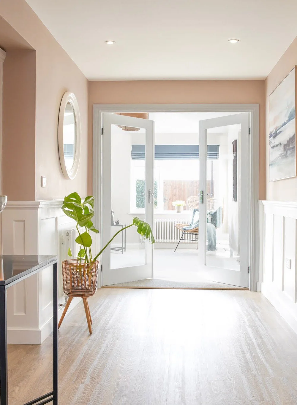 Bright entryway with open double glass doors leading to a sunlit sitting area with a chair, a throw blanket, and a large window with blinds.
