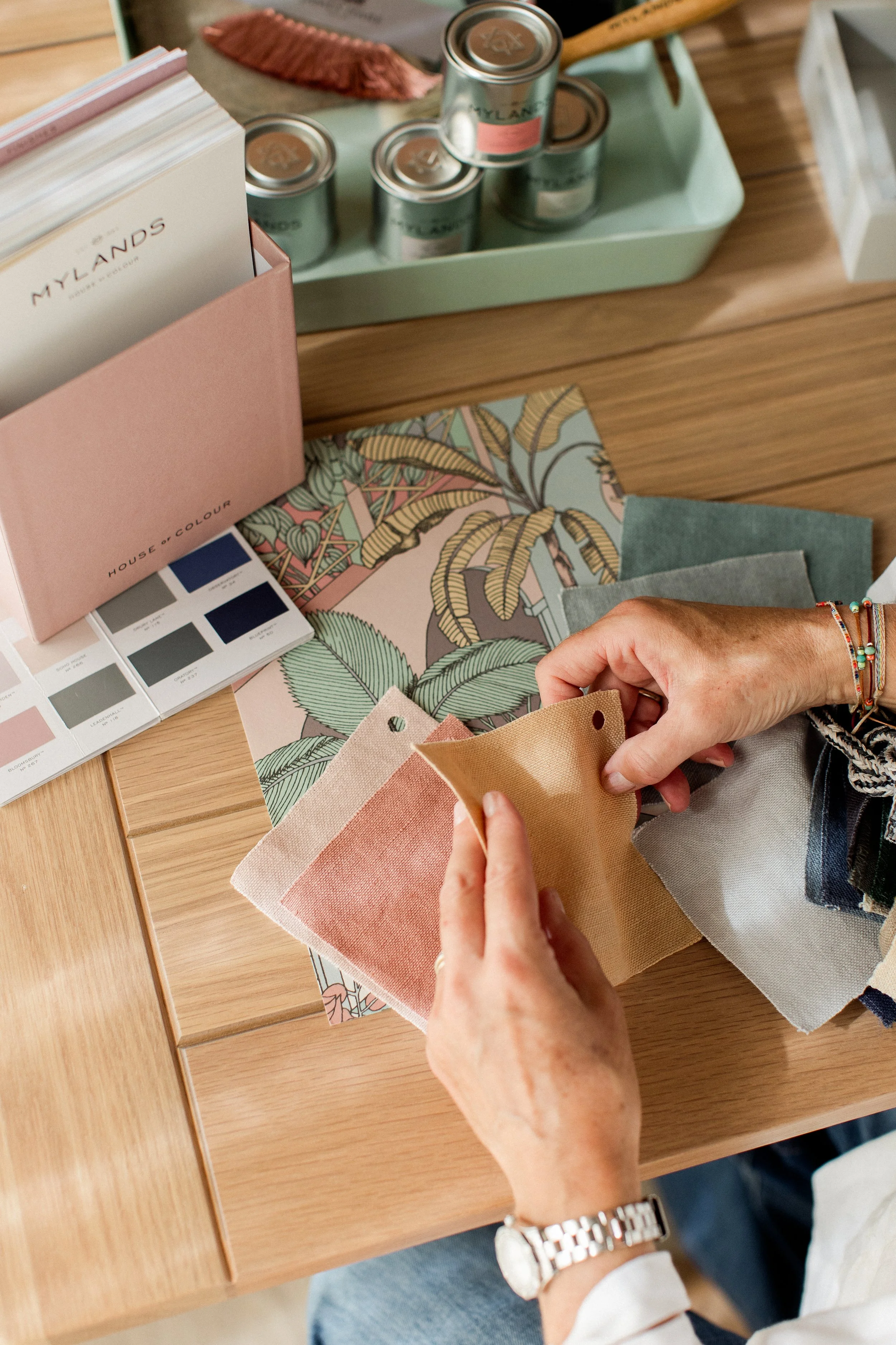 Person holding fabric swatches over a wooden table with color samples, a paint color palette, and various decorative items.