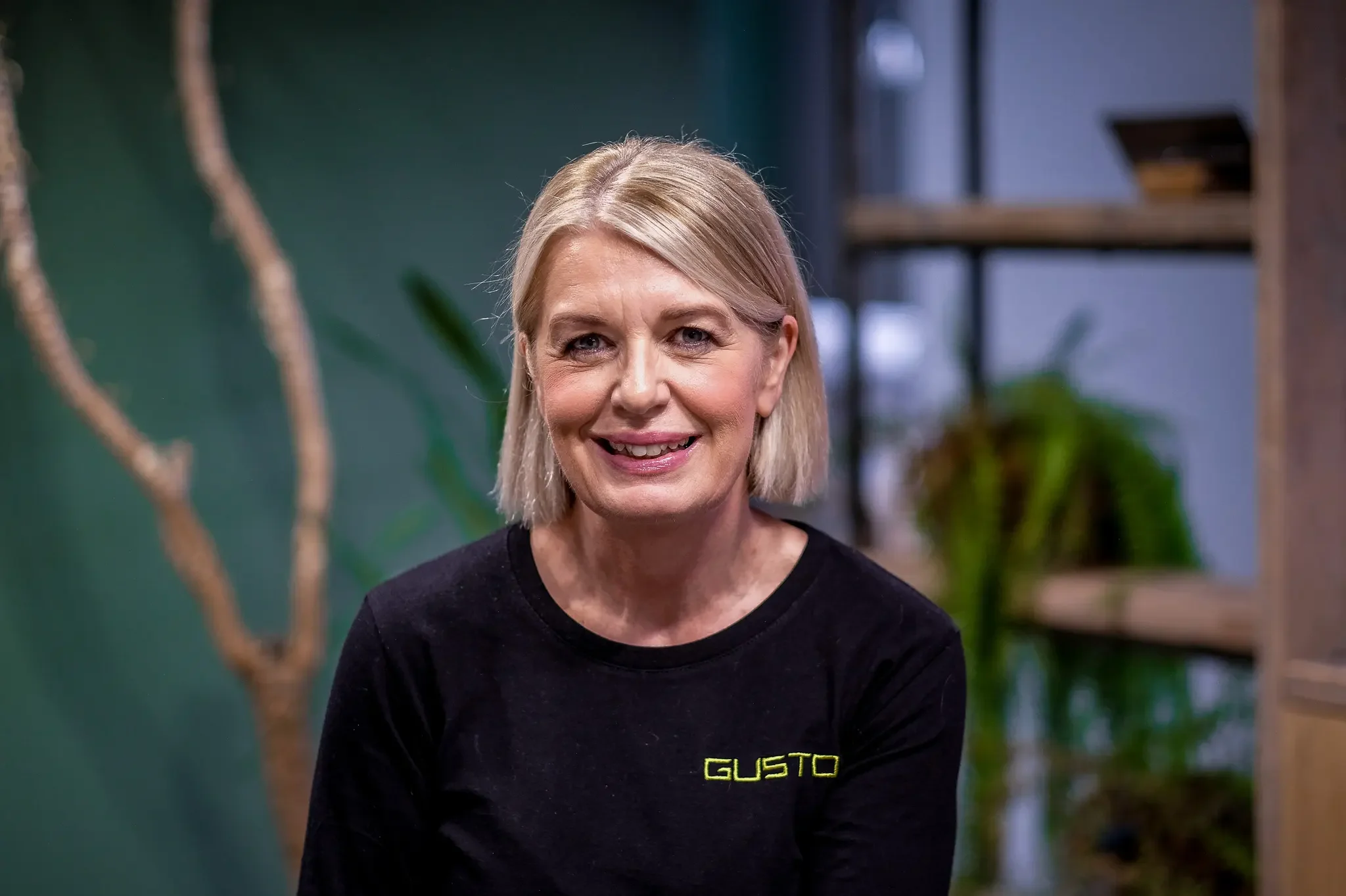 A middle aged women with short blond hair, pink lipstick, smiling in front of a green background wearing a black long sleeved top that says 'GUSTO' on it.