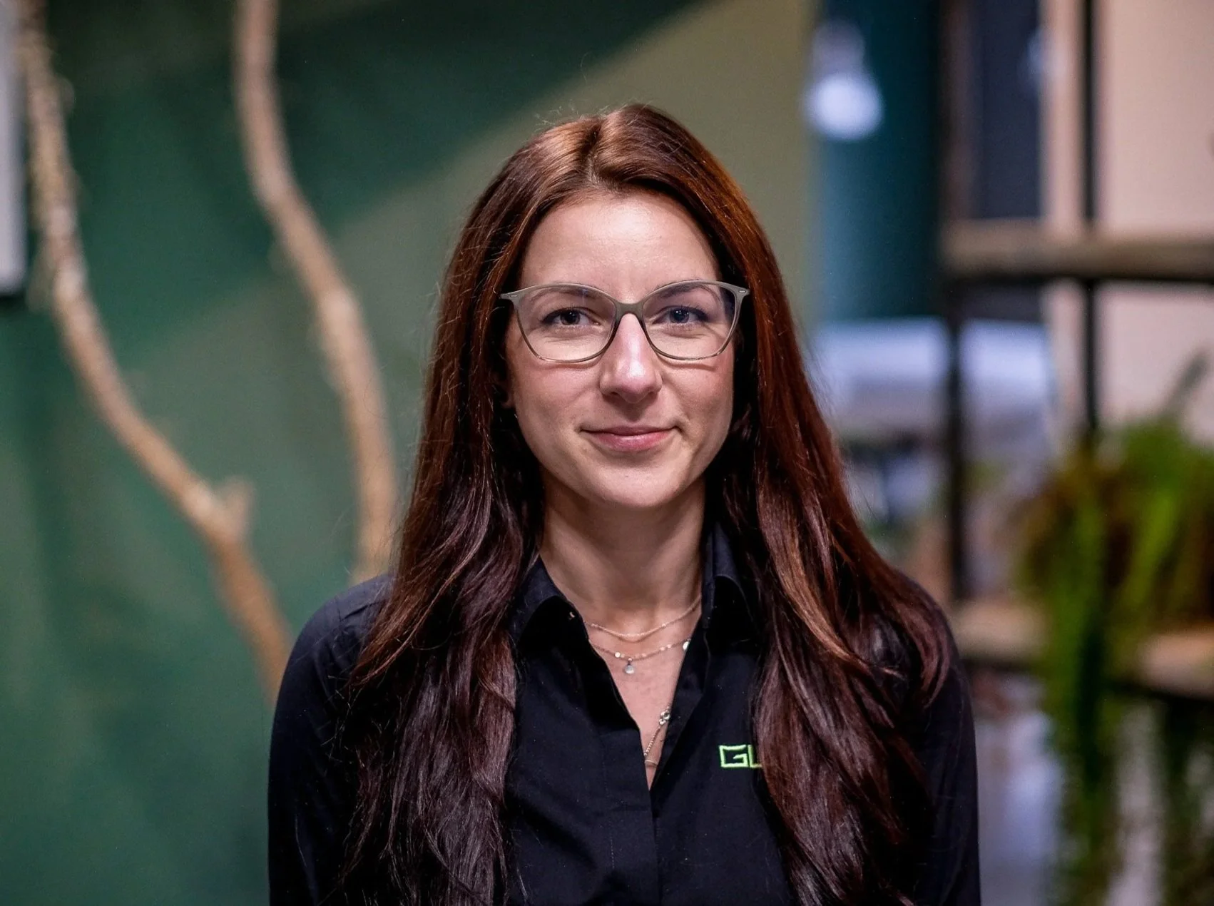 A woman with long dark hair, wearing glasses, silver earrings, and a necklace with a large triangular pendant. She is dressed in a black top and stands indoors against a green wall with a decorative branch to her left and a metal bookshelf with plants and lights in the background.