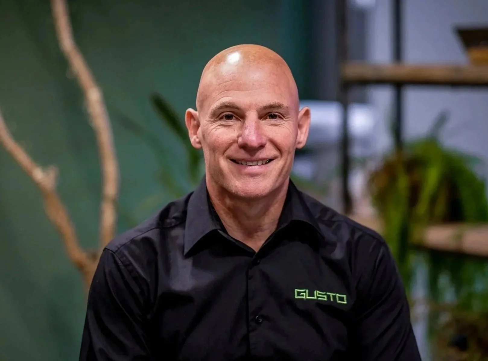 A middle-aged man with a shaved head and wearing a white button-down shirt, sitting indoors with a green wall and a potted plant in the background.
