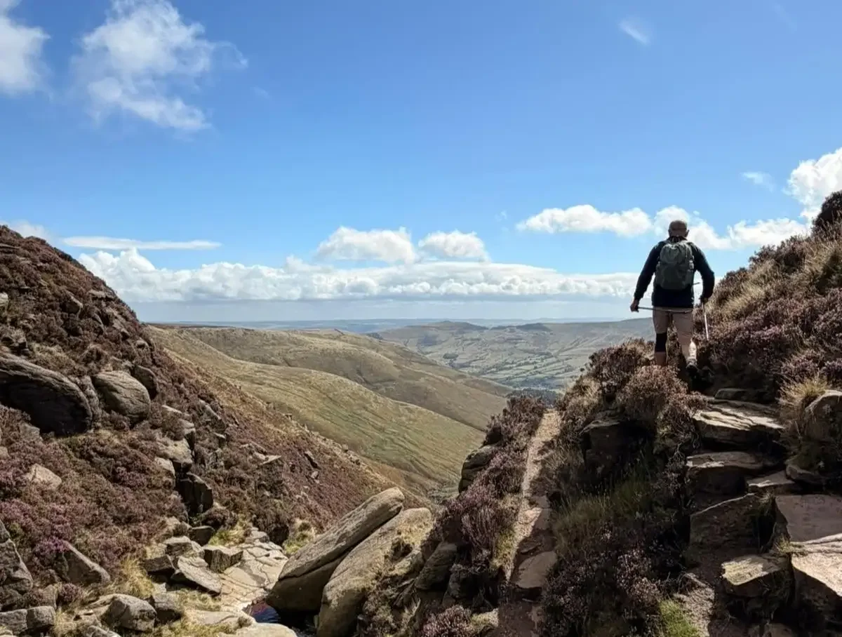A man walking up a mountain with a bagpack on with clouds and blue skies in the horizon.