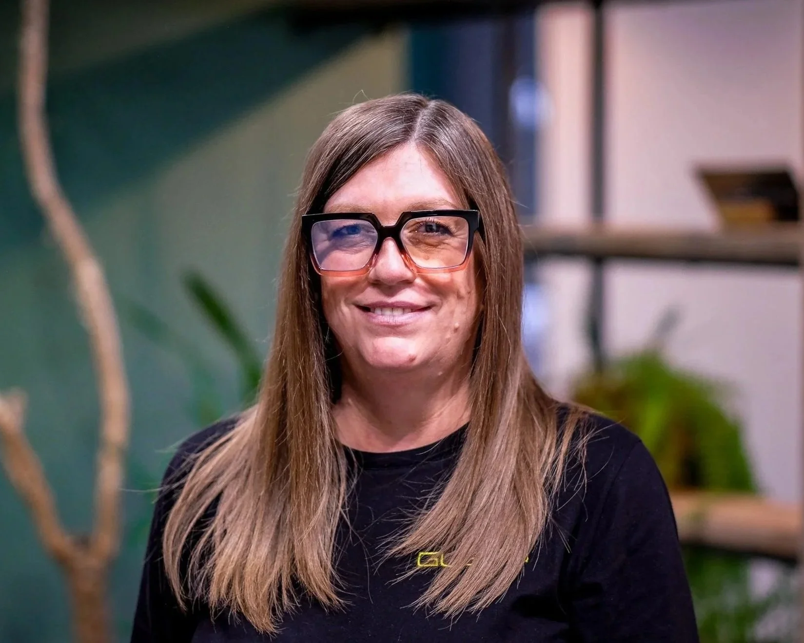 A smiling woman with long light brown hair wearing a black shirt, sitting indoors with a green wall and plants in the background.
