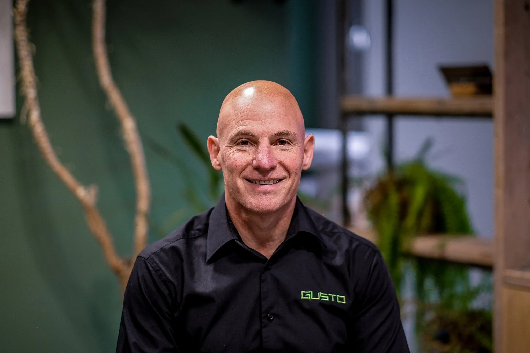 A middle-aged man with a shaved head and wearing a white button-down shirt, sitting indoors with a green wall and a potted plant in the background.