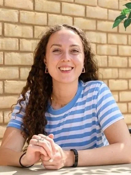 A young women with dark curly hair smiling in front of a brick wall