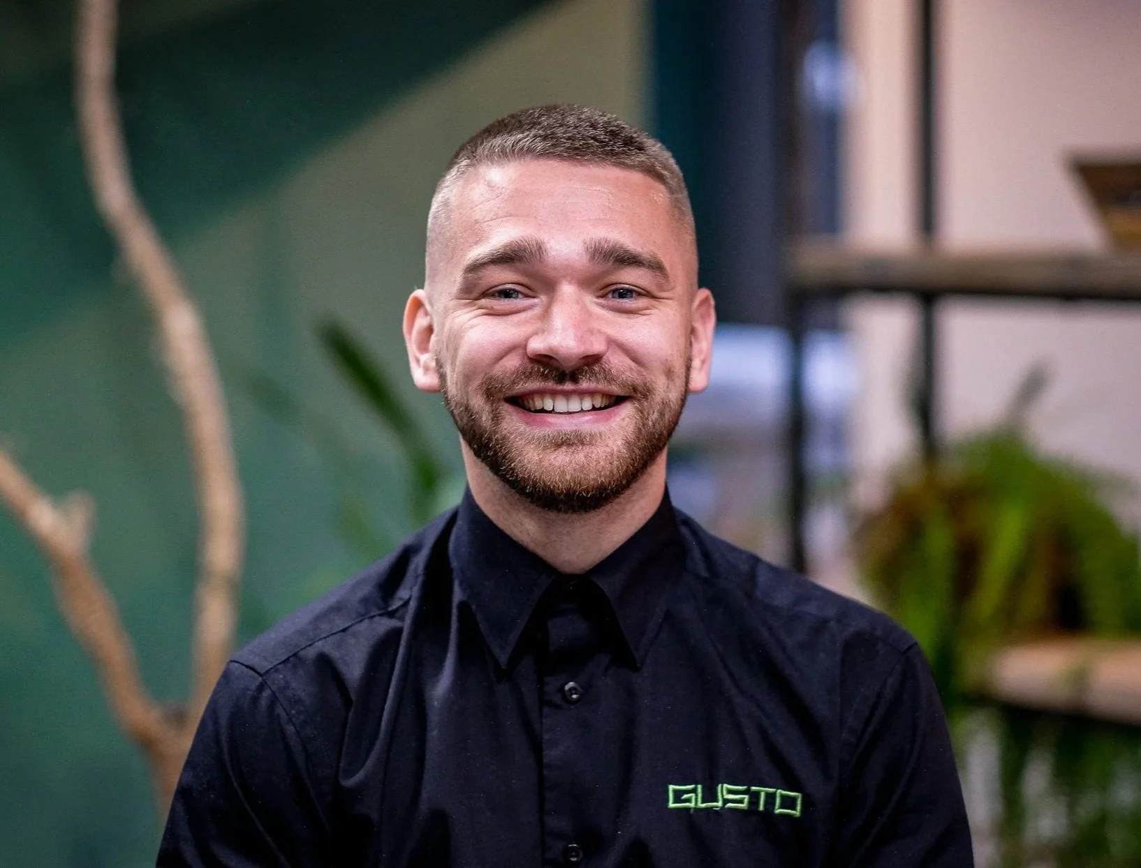 A smiling man with short brown hair, a beard, and blue eyes wearing a white collared shirt, in an office setting with a green wall and a blurred plant in the background.