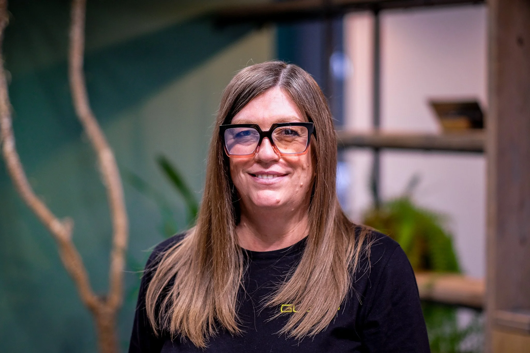 A smiling woman with long light brown hair wearing a black shirt, sitting indoors with a green wall and plants in the background.