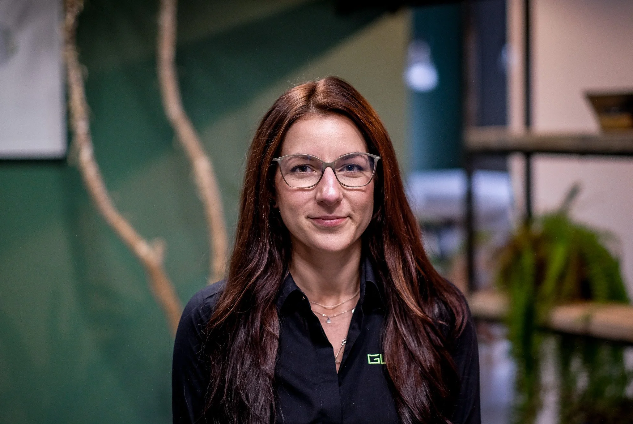 A woman with long dark hair, wearing glasses, silver earrings, and a necklace with a large triangular pendant. She is dressed in a black top and stands indoors against a green wall with a decorative branch to her left and a metal bookshelf with plants and lights in the background.