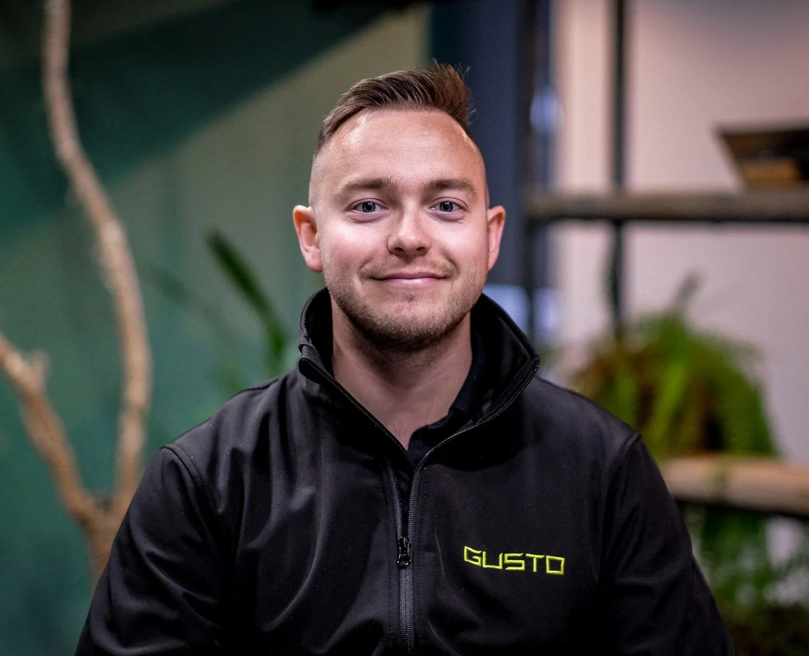 A young man with short brown hair, wearing glasses and a black zip-up sweater, smiling indoors against a green wall and surrounding plants.