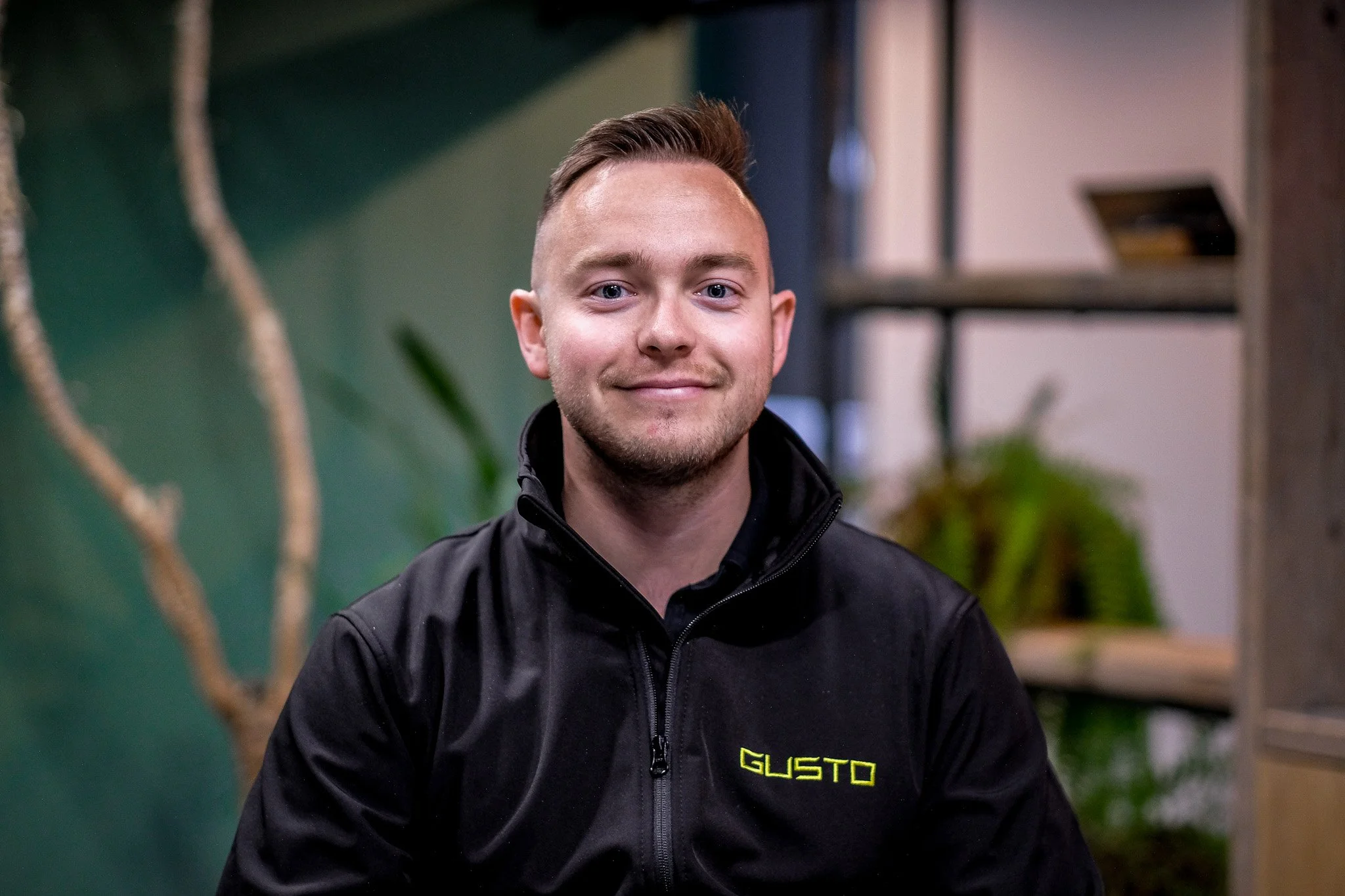 A young man with short brown hair, wearing glasses and a black zip-up sweater, smiling indoors against a green wall and surrounding plants.