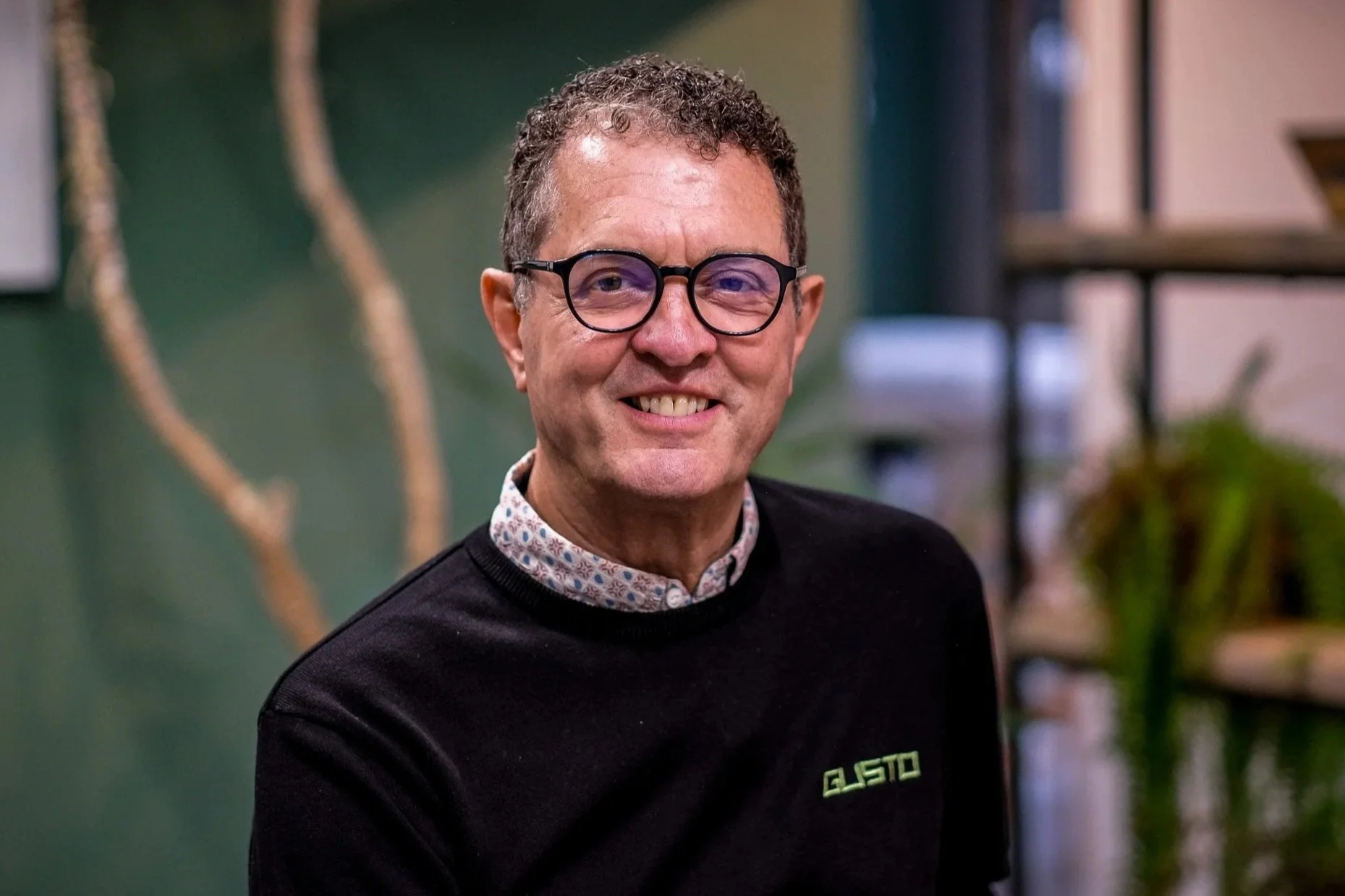 A middle-aged man with curly brown hair, wearing glasses and a checkered shirt, looking at the camera with a slight smile, standing in front of a blurred background with green and neutral tones.