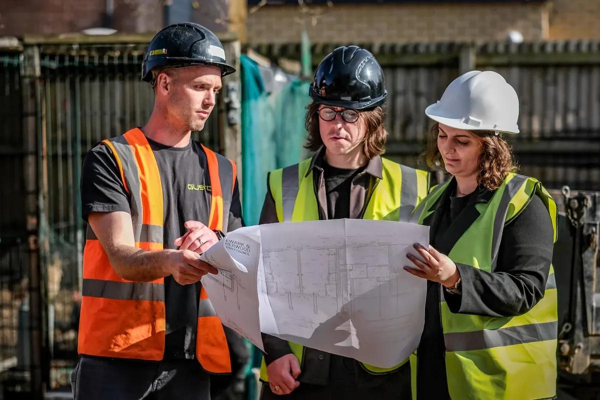 a construction worker and two architects on site, wearing hi vis and hard hats, looking at a drawing/site plans