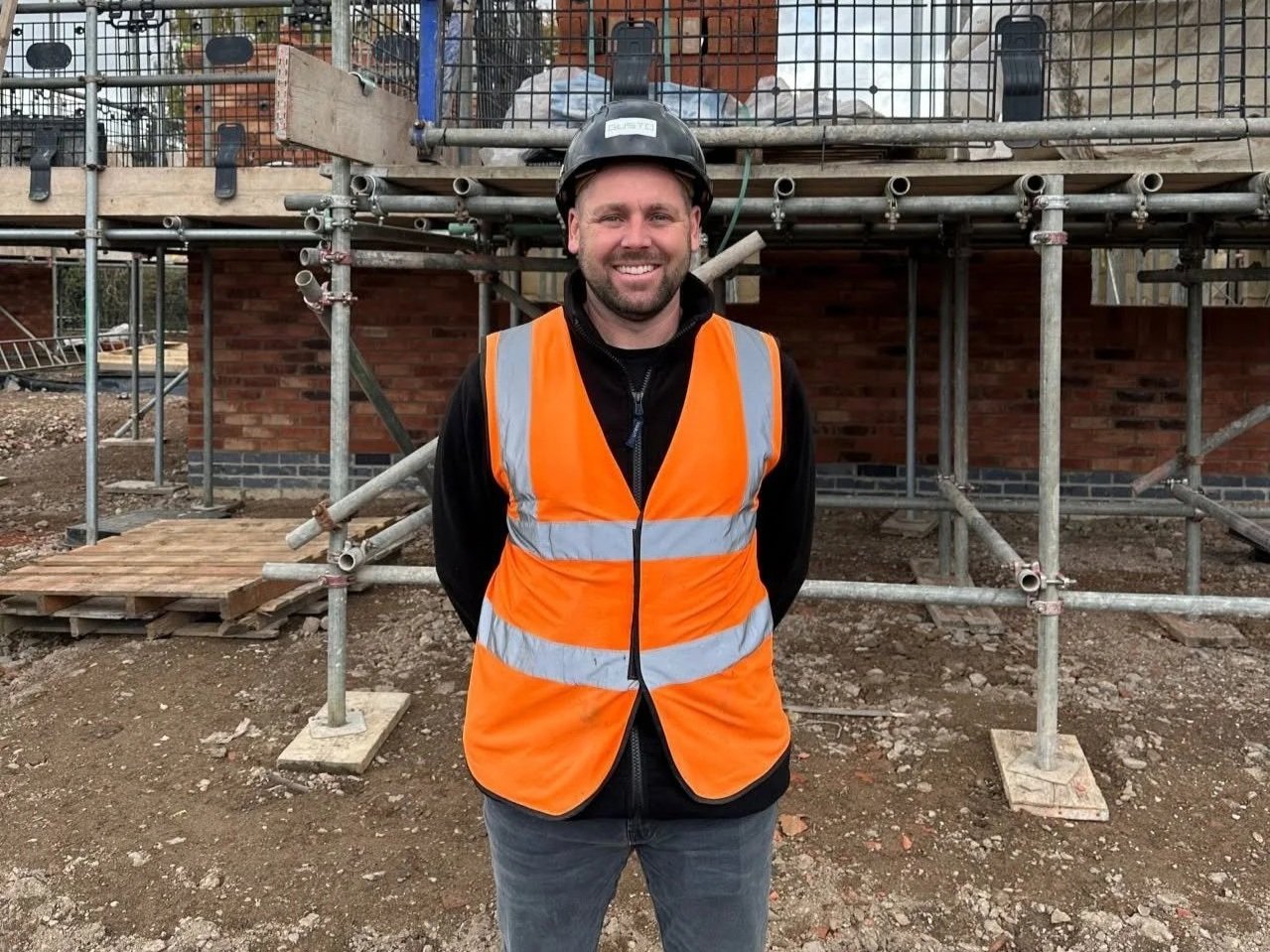 A Site Manager from Gusto Construction on a construction site, stood in front of a house being built, in a black hard hat that says Gusto and an orange hi-vis vest.