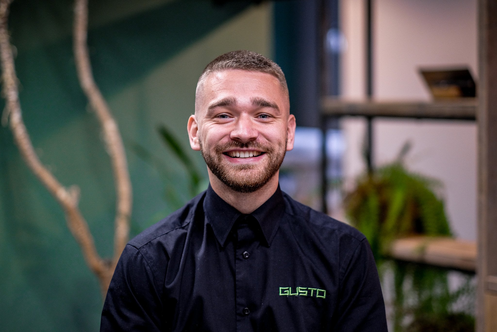 A smiling man with short brown hair, a beard, and blue eyes wearing a white collared shirt, in an office setting with a green wall and a blurred plant in the background.