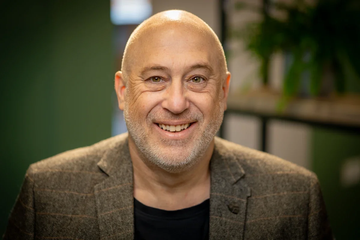 A bald man with stubble, wearing a tweed jacket and smiling at the camera in front of a green wall and indoor plants