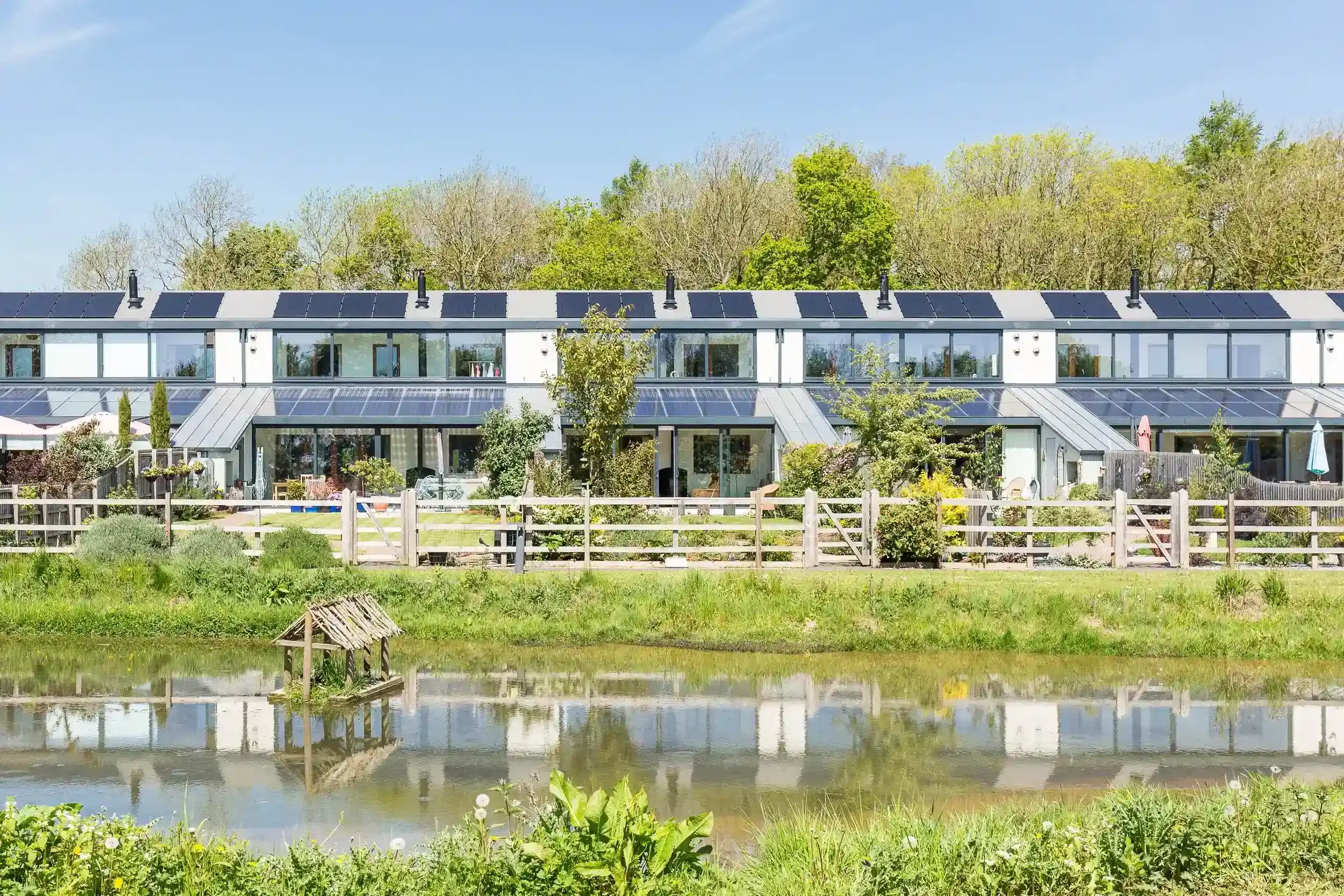 Row of modern house with solar panels on roof, surrounded by trees and garden, beside a pond with a small wooden structure.