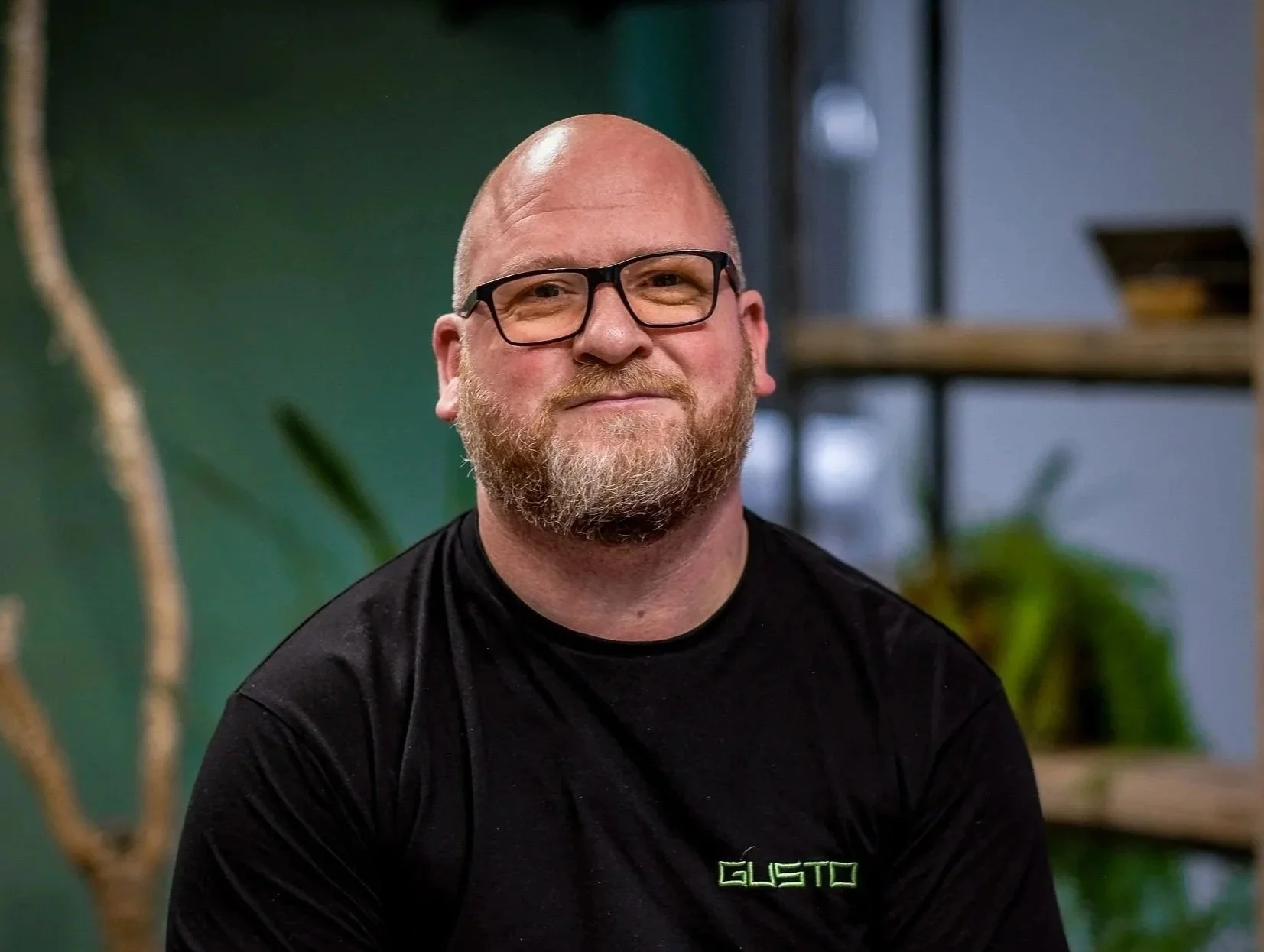 A bald man with glasses and a beard, wearing a black shirt with the logo 'GUSTO', smiling at the camera indoors with a green wall, wooden shelves, potted plants, and blurred background.