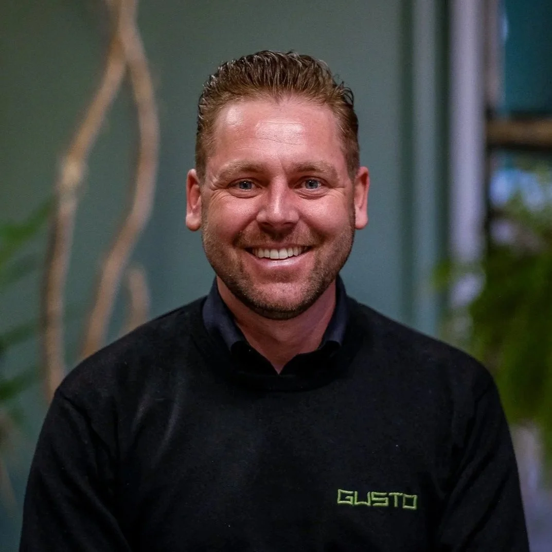 A smiling man with short blond hair and a beard, wearing a black jacket over a dark blue sweater, standing indoors with green walls and plants in the background.